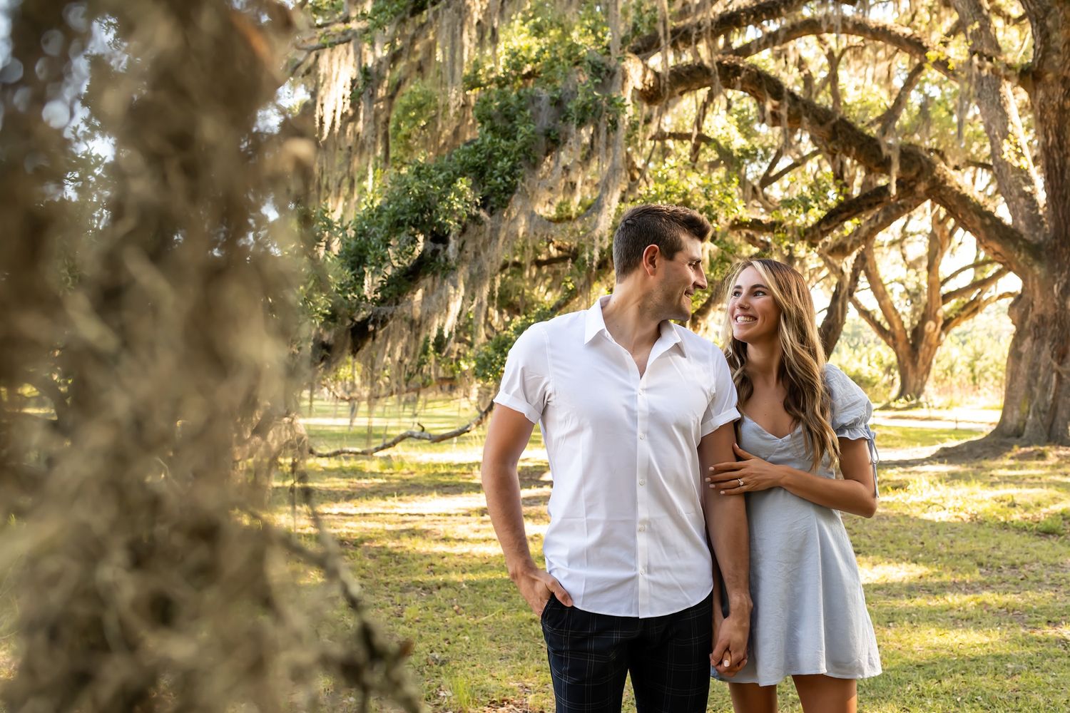 A couple shares a tender moment while walking together under moss-draped oak trees in a sun-dappled park.