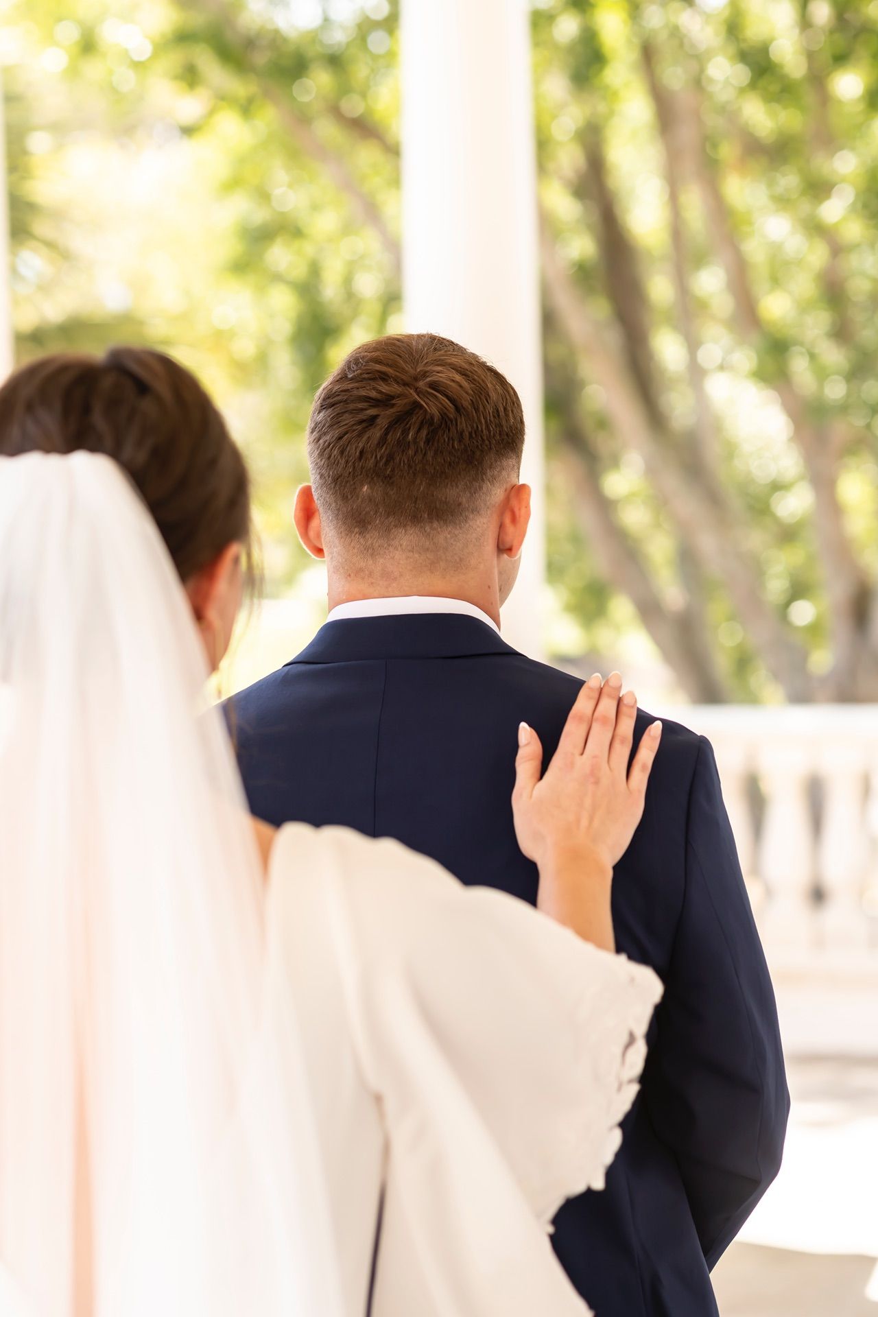 Groomsmen in navy suits stand in a sun-dappled outdoor setting during a wedding ceremony.