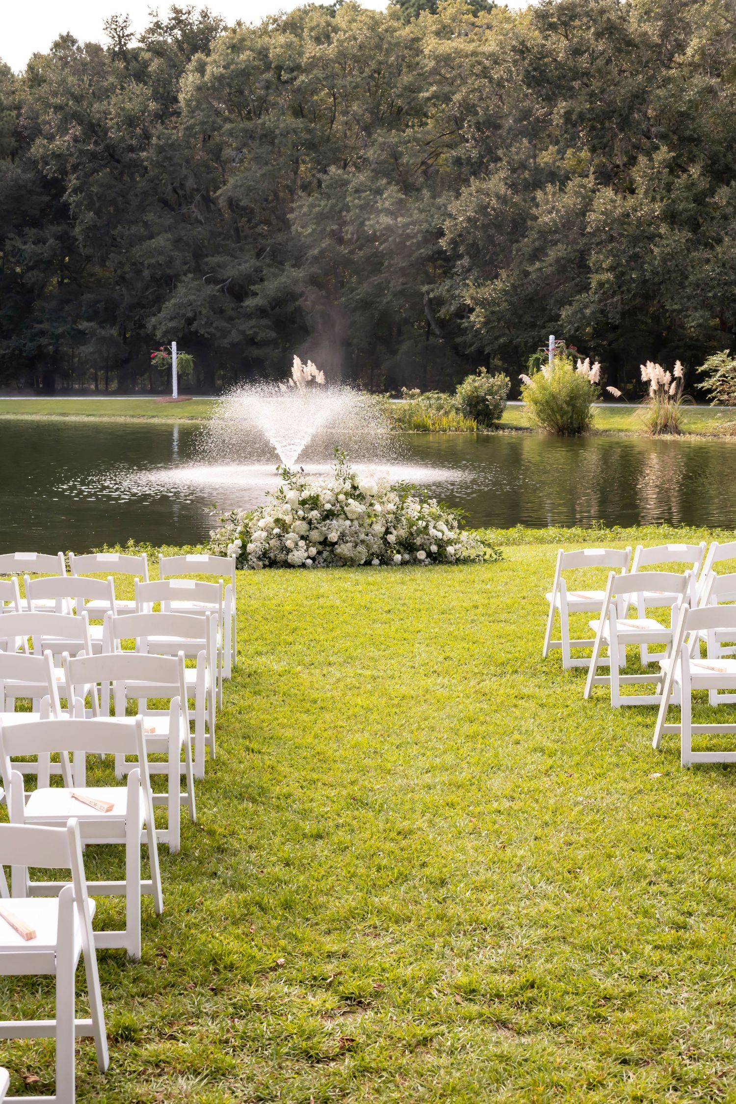 Outdoor wedding ceremony setup with white chairs on green lawn, floral arrangement, and fountain by a serene lake backdrop.
