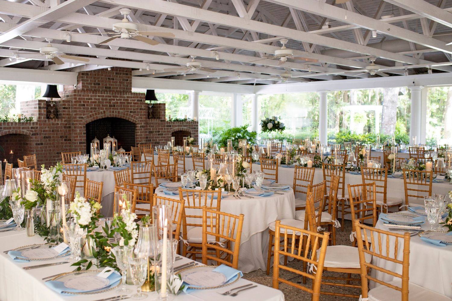 Elegant wedding reception hall with gold Chiavari chairs, white tablecloths, floral centerpieces, and a brick fireplace.