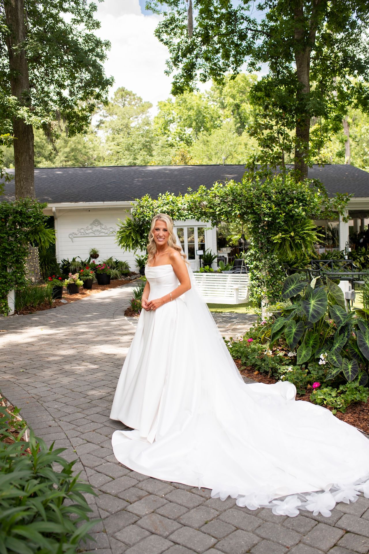 Bride in strapless white ball gown with long train poses on brick path in lush garden setting.