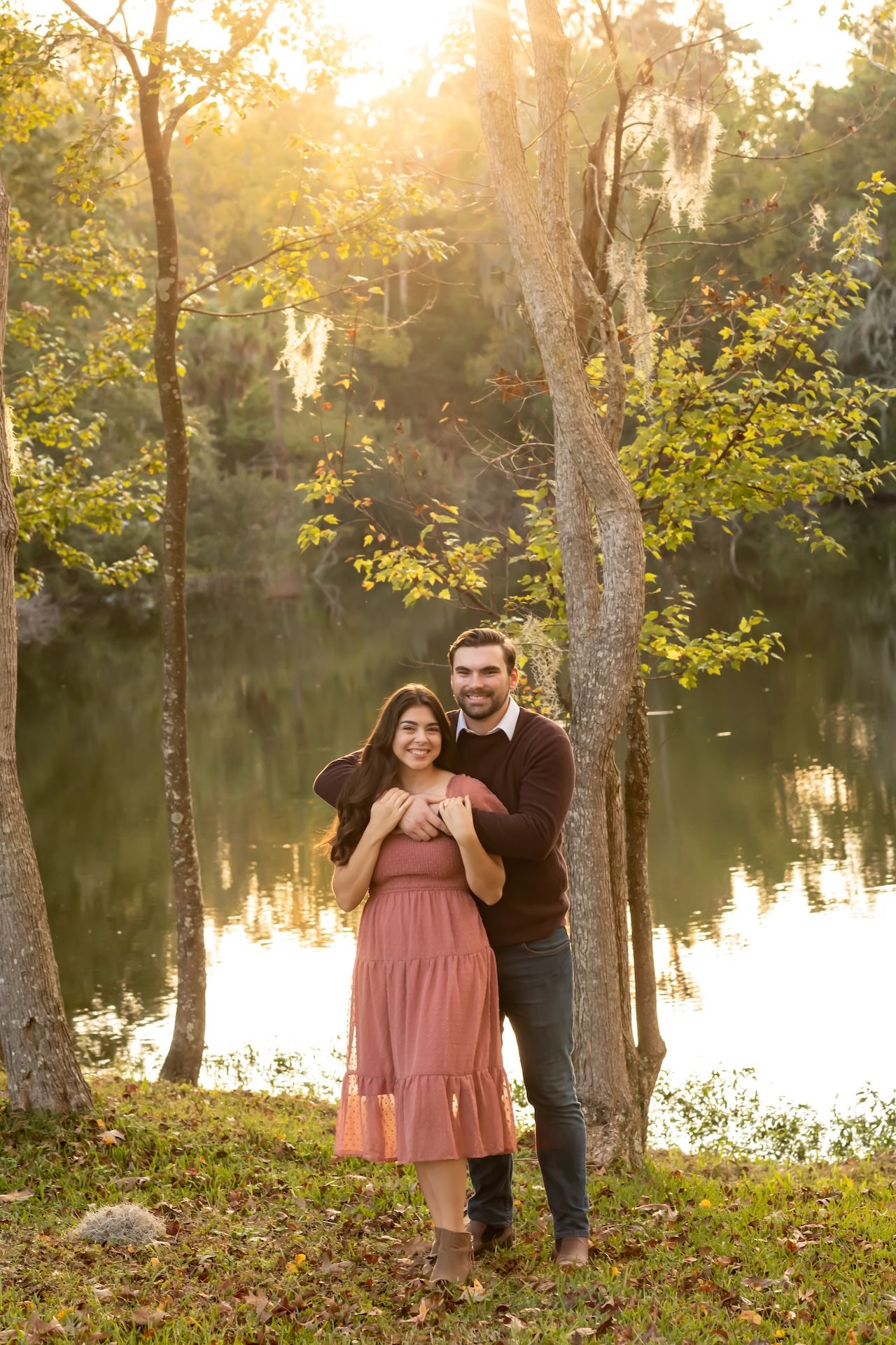Couple posing by a sunlit lake surrounded by trees in golden hour light, woman in pink dress, man in dark sweater.