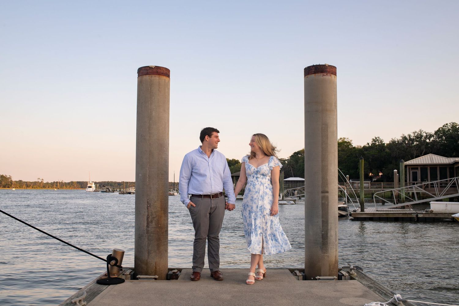 A romantic couple stands between pillars at a waterfront dock during sunset with boats in the background.