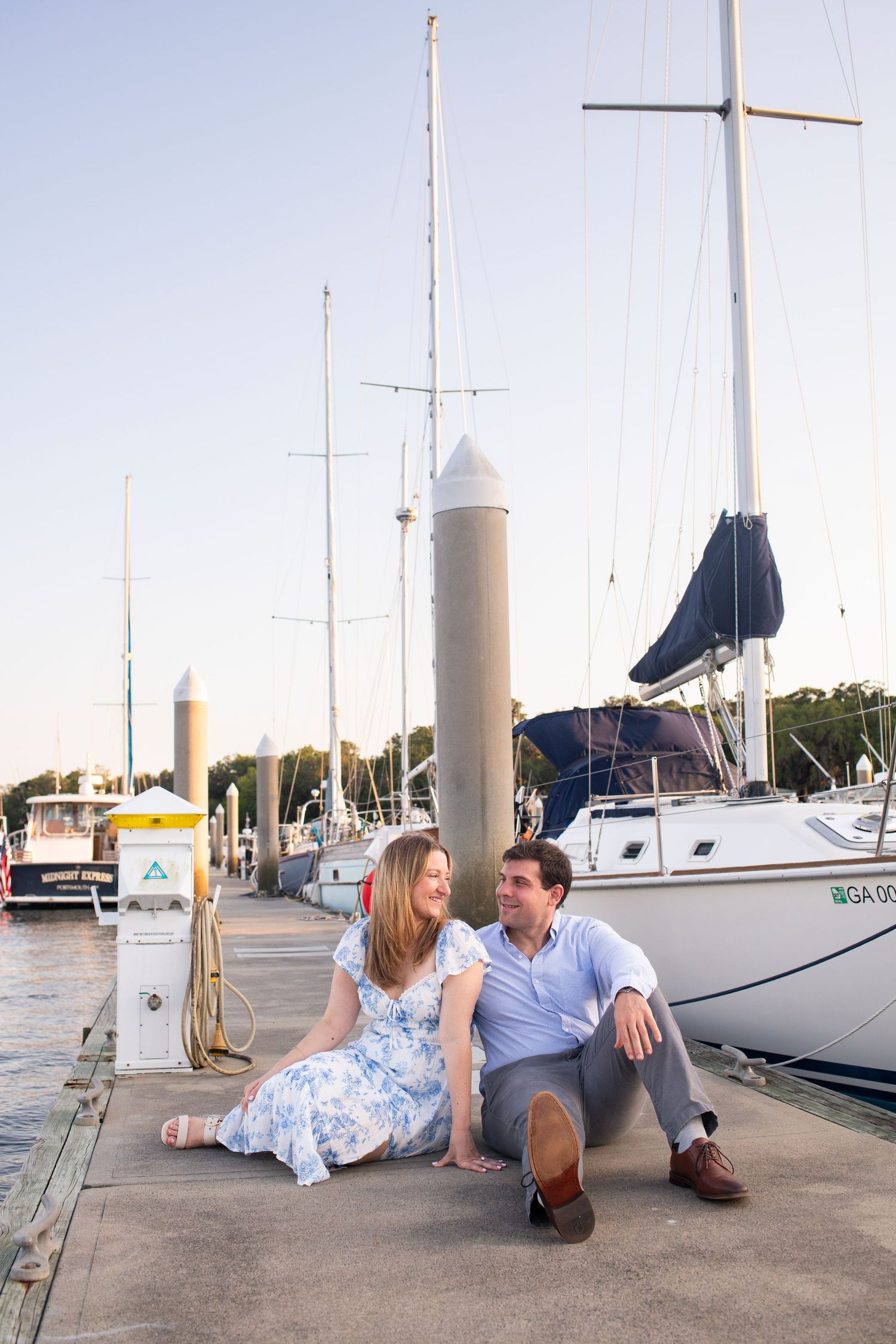 A romantic couple sits together on a marina dock with sailboats in the background during golden hour.