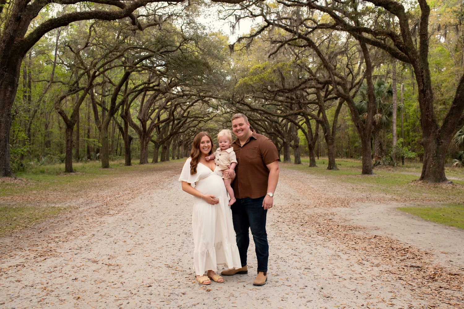 Pregnant couple with toddler son poses on tree-lined path for maternity photo session outdoors.