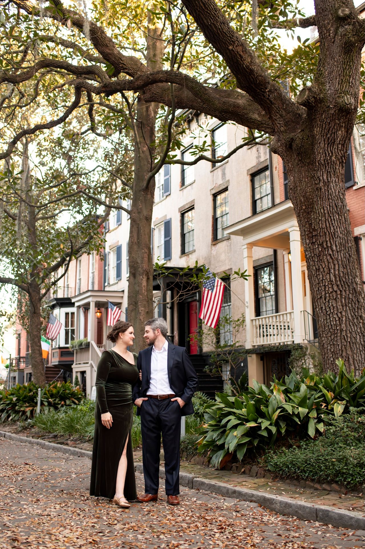Historic brownstone buildings with American flags line a tree-shaded sidewalk in Savannah's historic district.