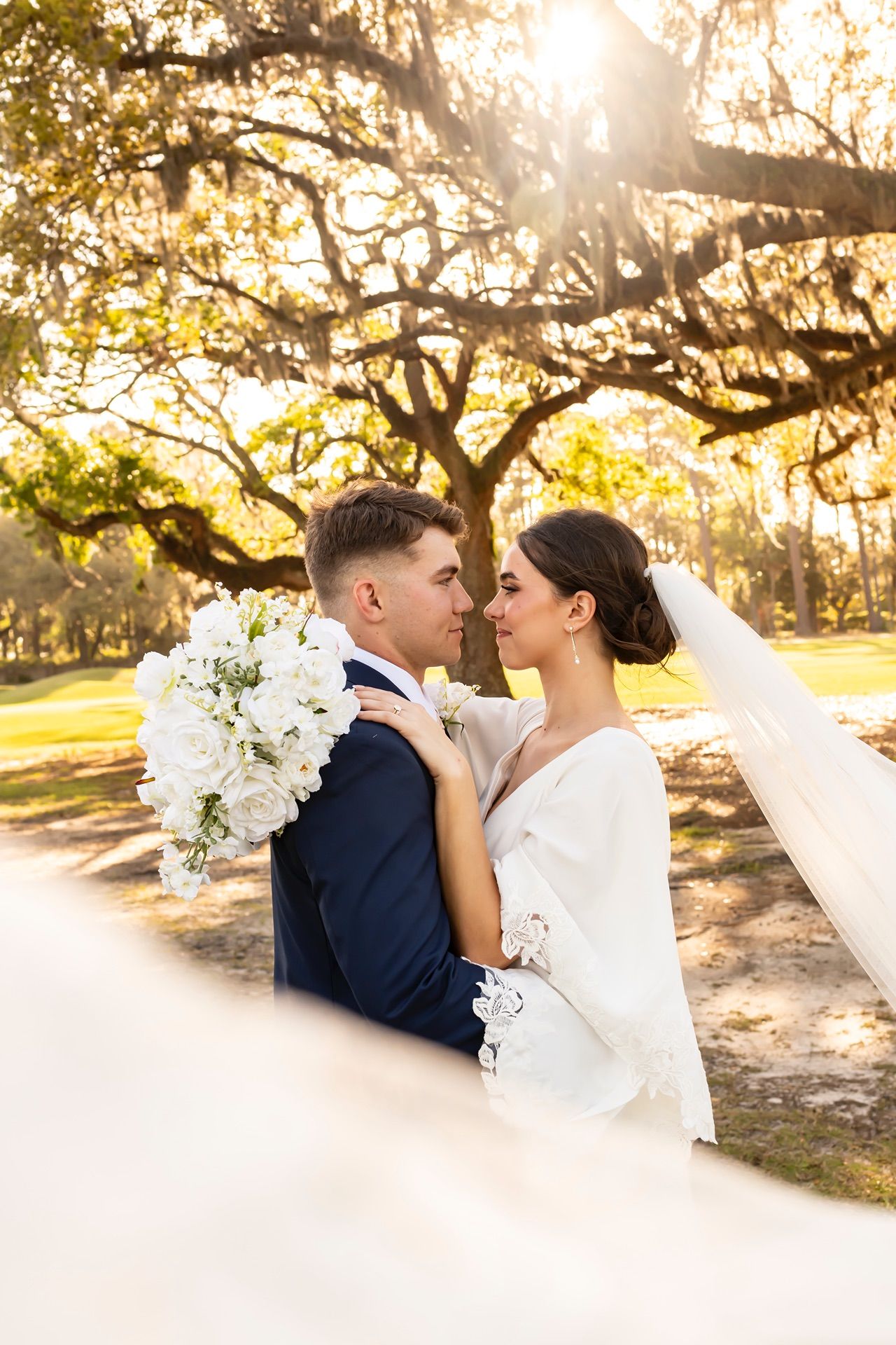 A romantic wedding couple embraces under golden sunlit oak trees in a dreamy outdoor setting.