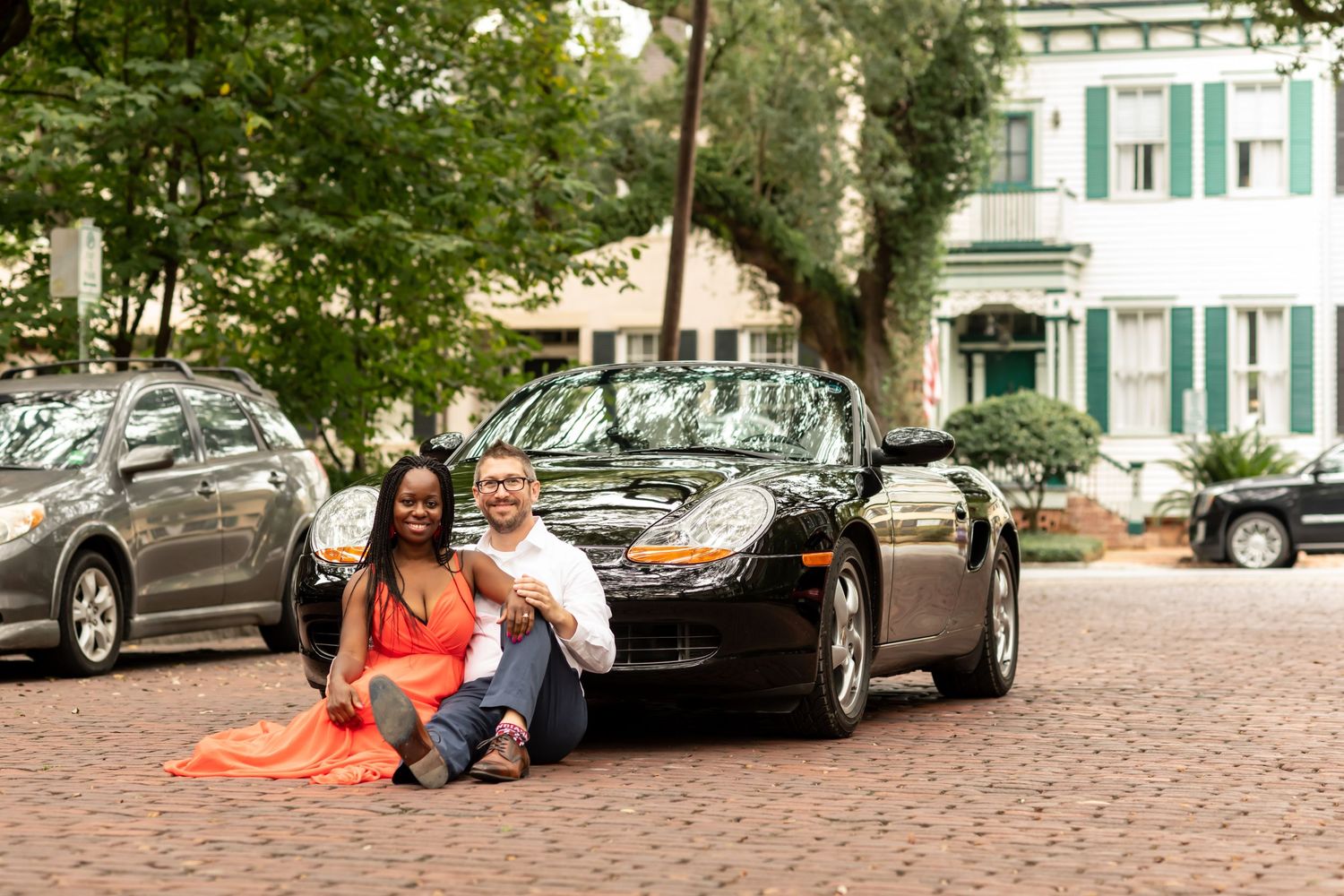 A couple sits together on the hood of a black car parked on a cobblestone street near historic buildings.