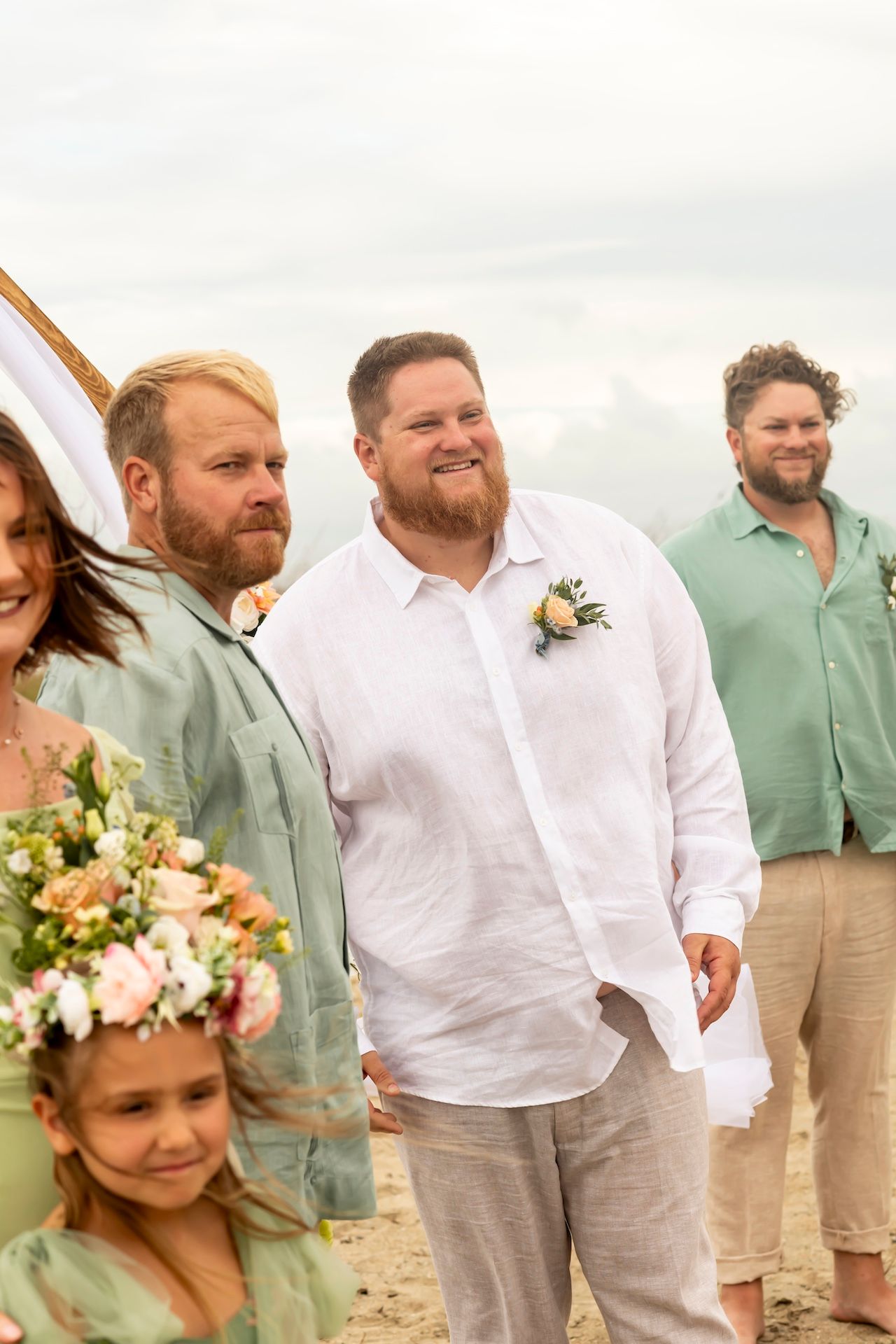Wedding party members in sage green and white linen attire smile together on a sunny beach.
