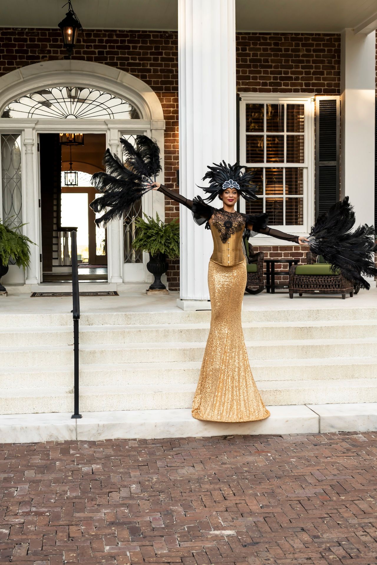 Golden dress sculpture with black feathered wings stands on a brick porch against white columns and windows.
