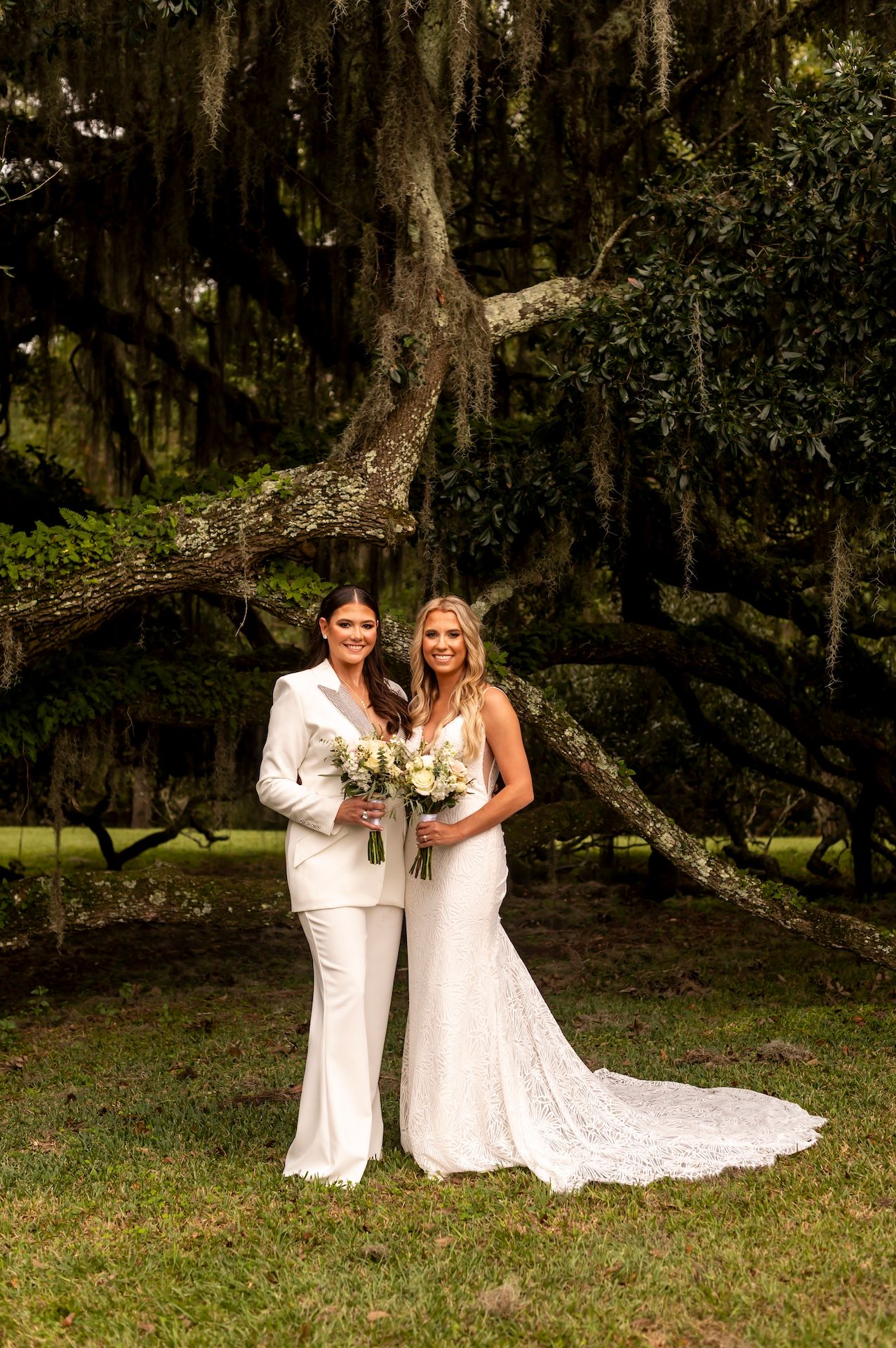 A wedding couple poses together beneath Spanish moss draped oak trees in a romantic Southern garden setting.