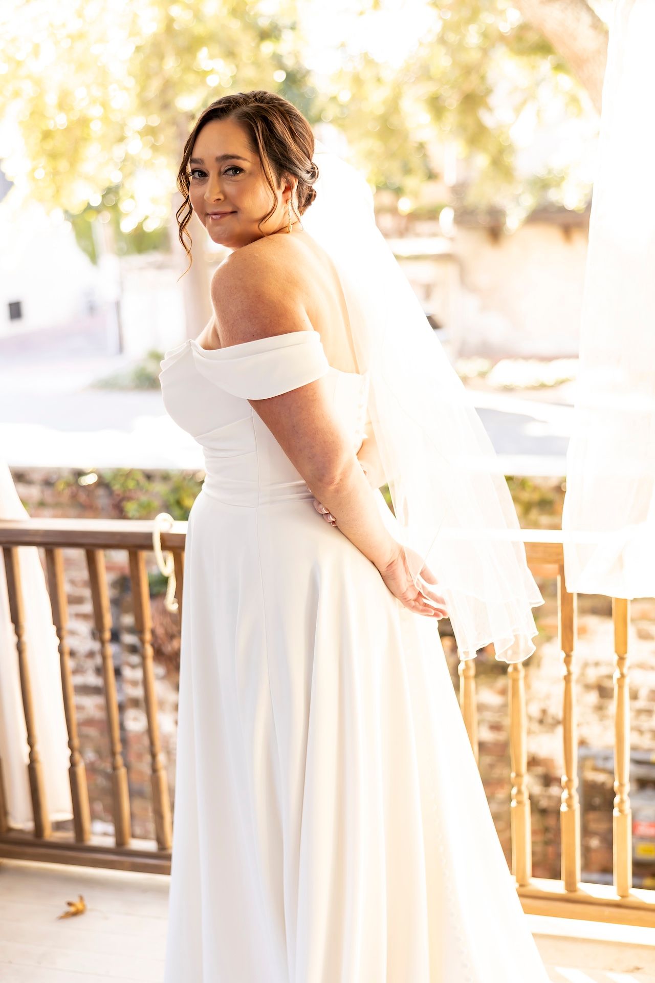 A bride in an elegant white off-shoulder wedding gown poses gracefully on a sunlit outdoor deck.