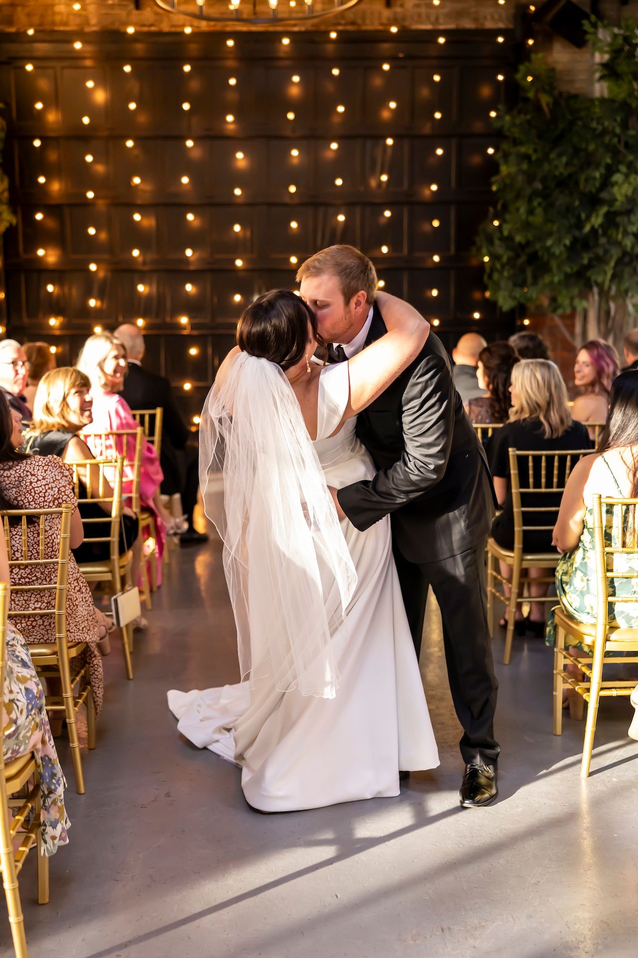 A couple shares their first dance at a reception with twinkle lights and gold chairs creating a romantic atmosphere.