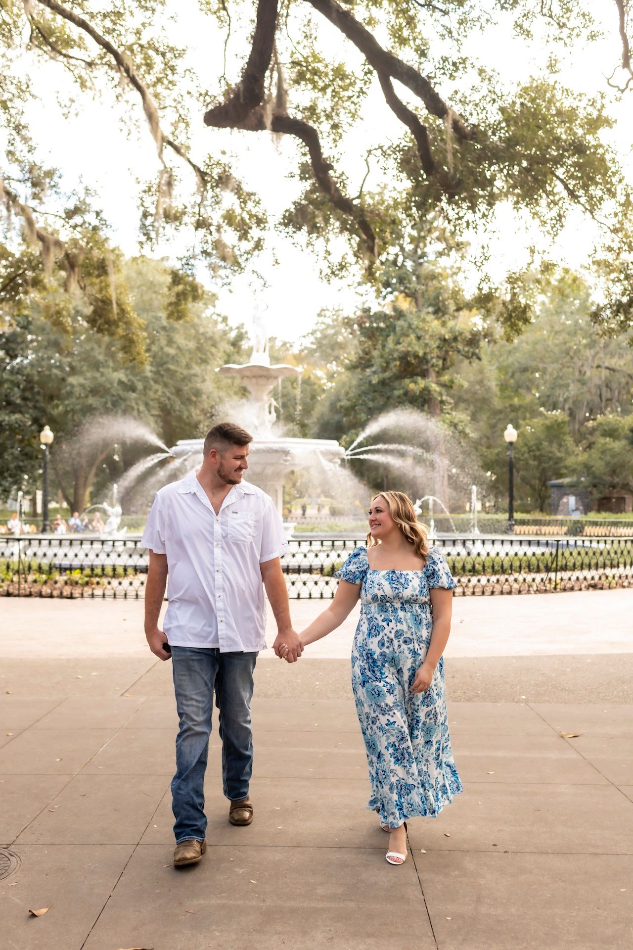 A couple walks hand in hand on a paved pathway with a scenic fountain and lush trees in the background.