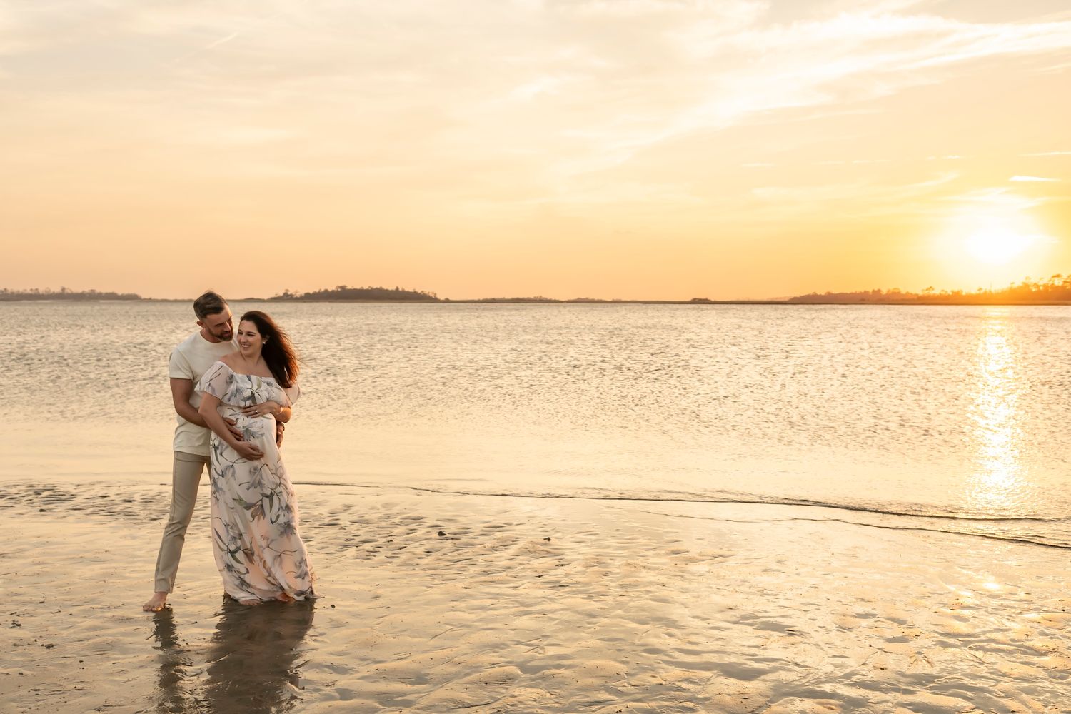 Romantic couple embraces while standing in shallow water during golden sunset on tropical beach.