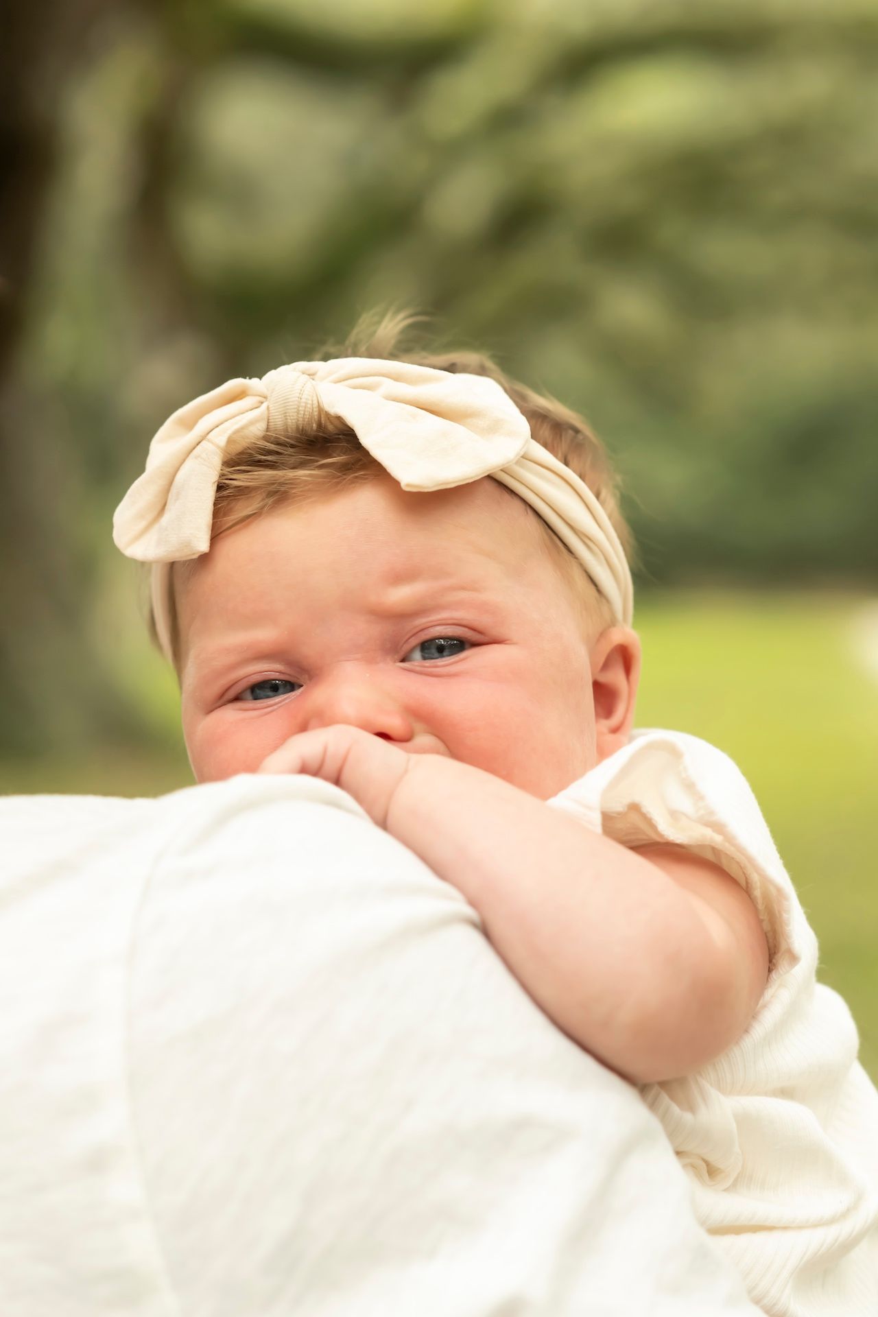 Baby wearing cream-colored outfit and headband smiles while snuggling against white fabric outdoors.