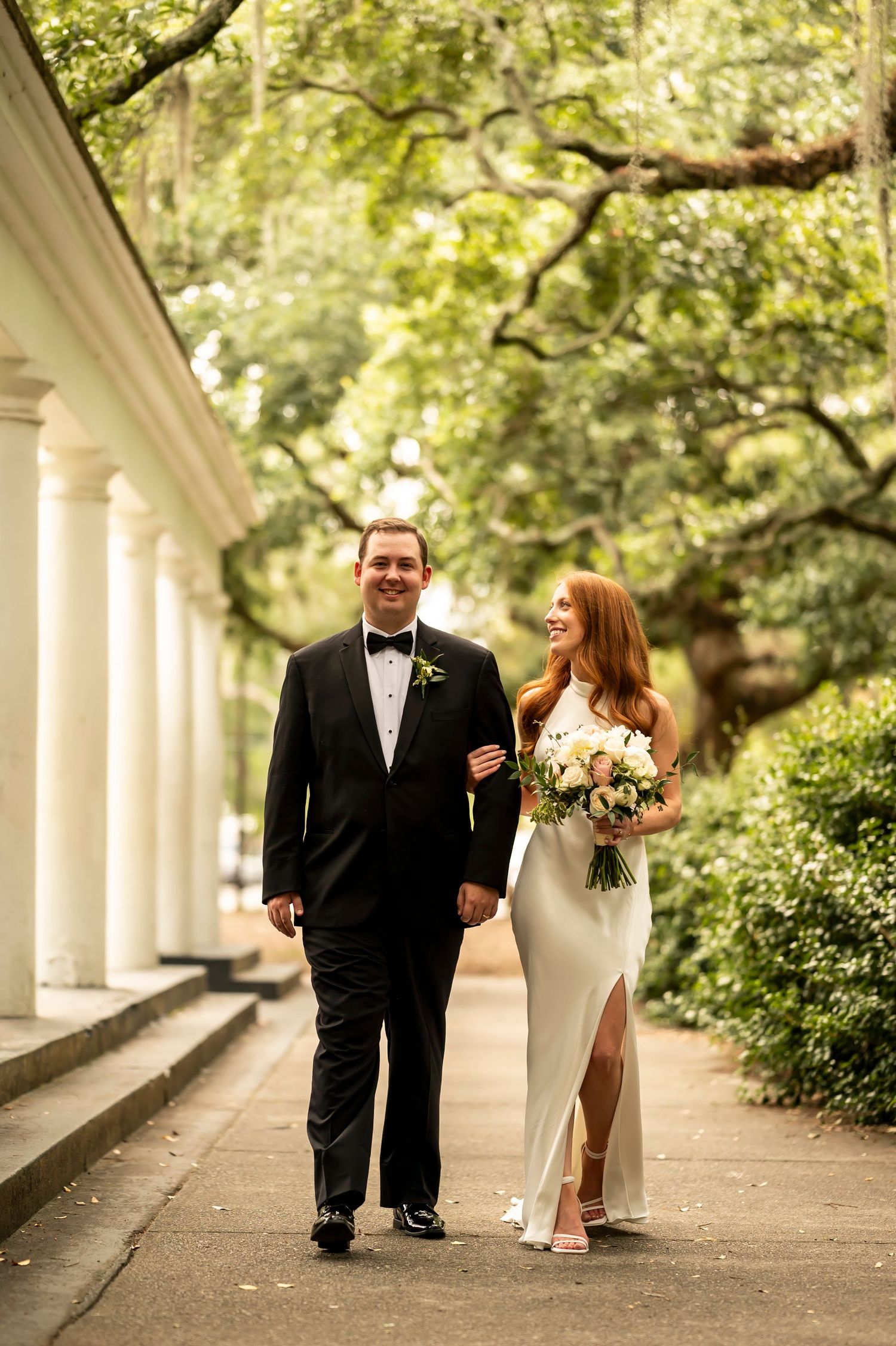 Newlywed couple walking arm in arm along a columned pathway surrounded by lush greenery and trees.
