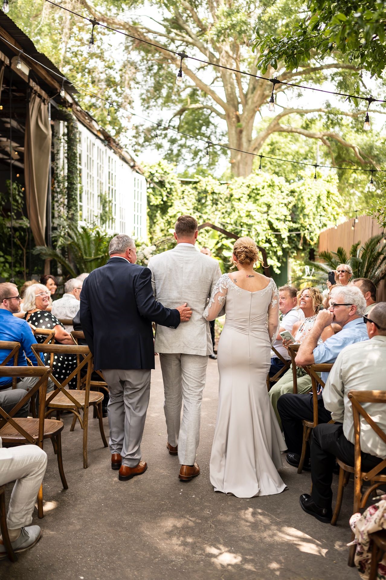 Couple walks down outdoor venue aisle during wedding ceremony with guests seated on wooden chairs in garden setting.