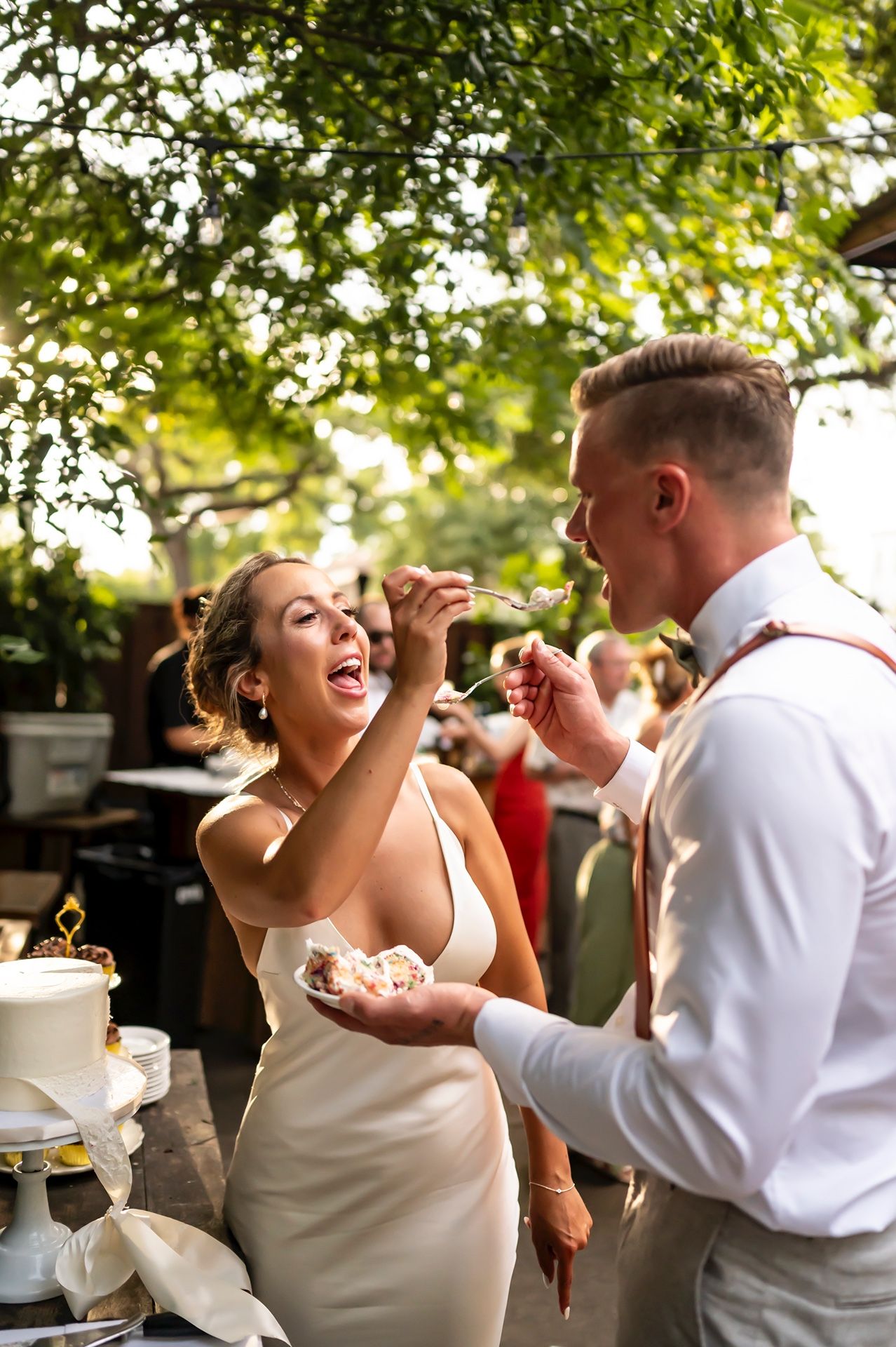 Couple playfully feeds each other wedding cake during outdoor reception ceremony under green tree canopy.