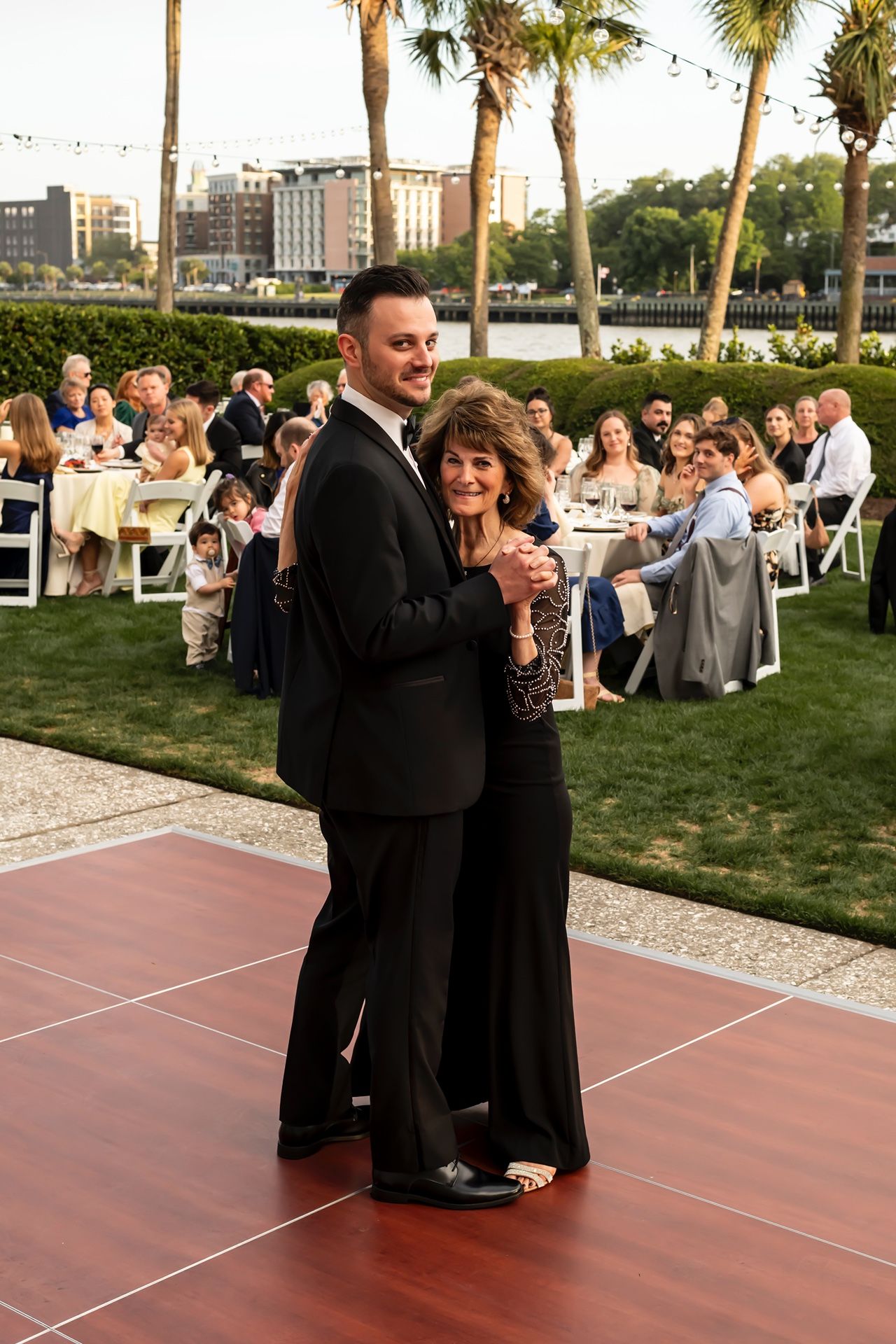 Guests dance on an outdoor dance floor during a sunset wedding reception with palm trees and city buildings in the background.
