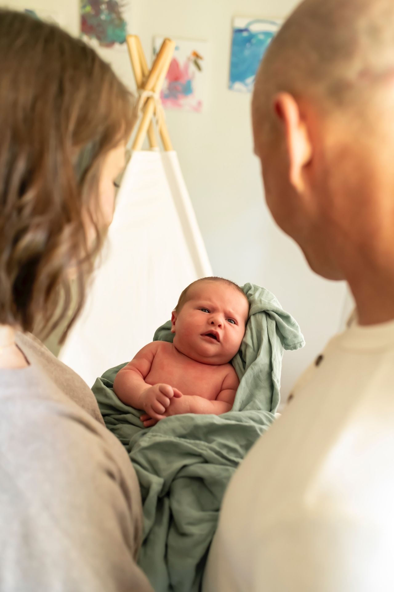 A newborn baby wrapped in a green blanket lies on a surface while two adults look down at them in a room with a teepee.
