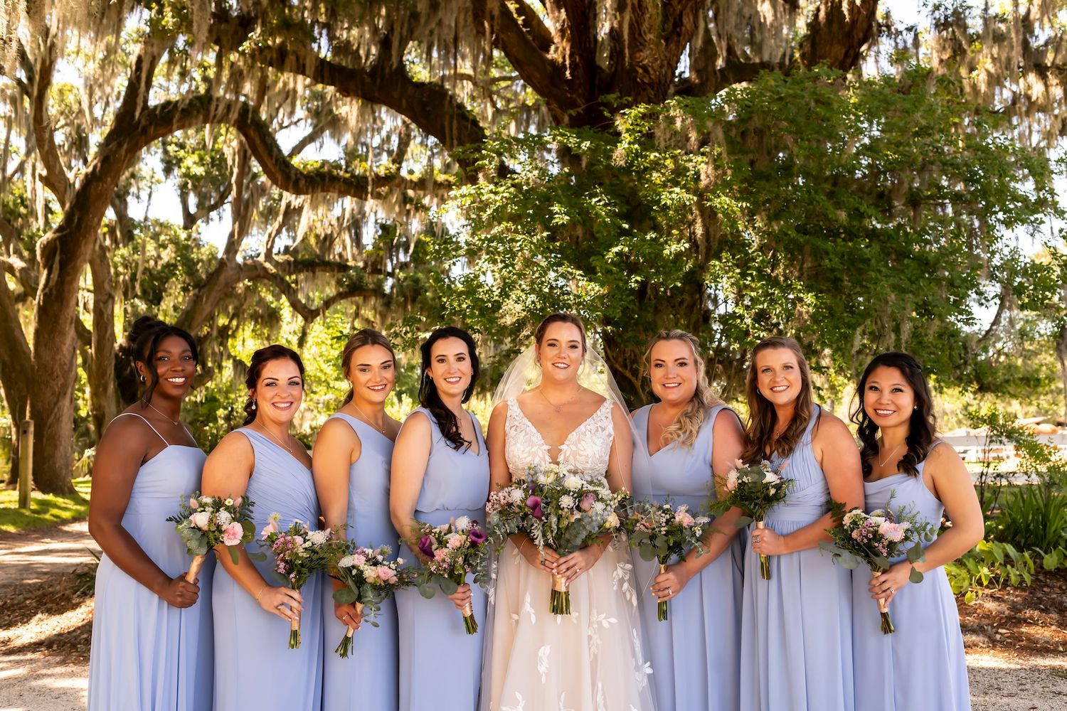A group poses in light blue bridesmaid dresses and white wedding gown under majestic oak trees with Spanish moss.