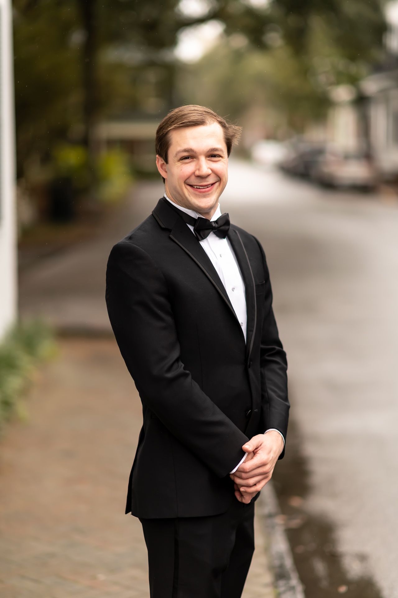 A person in a classic black tuxedo and bow tie smiles while standing on a wet sidewalk.