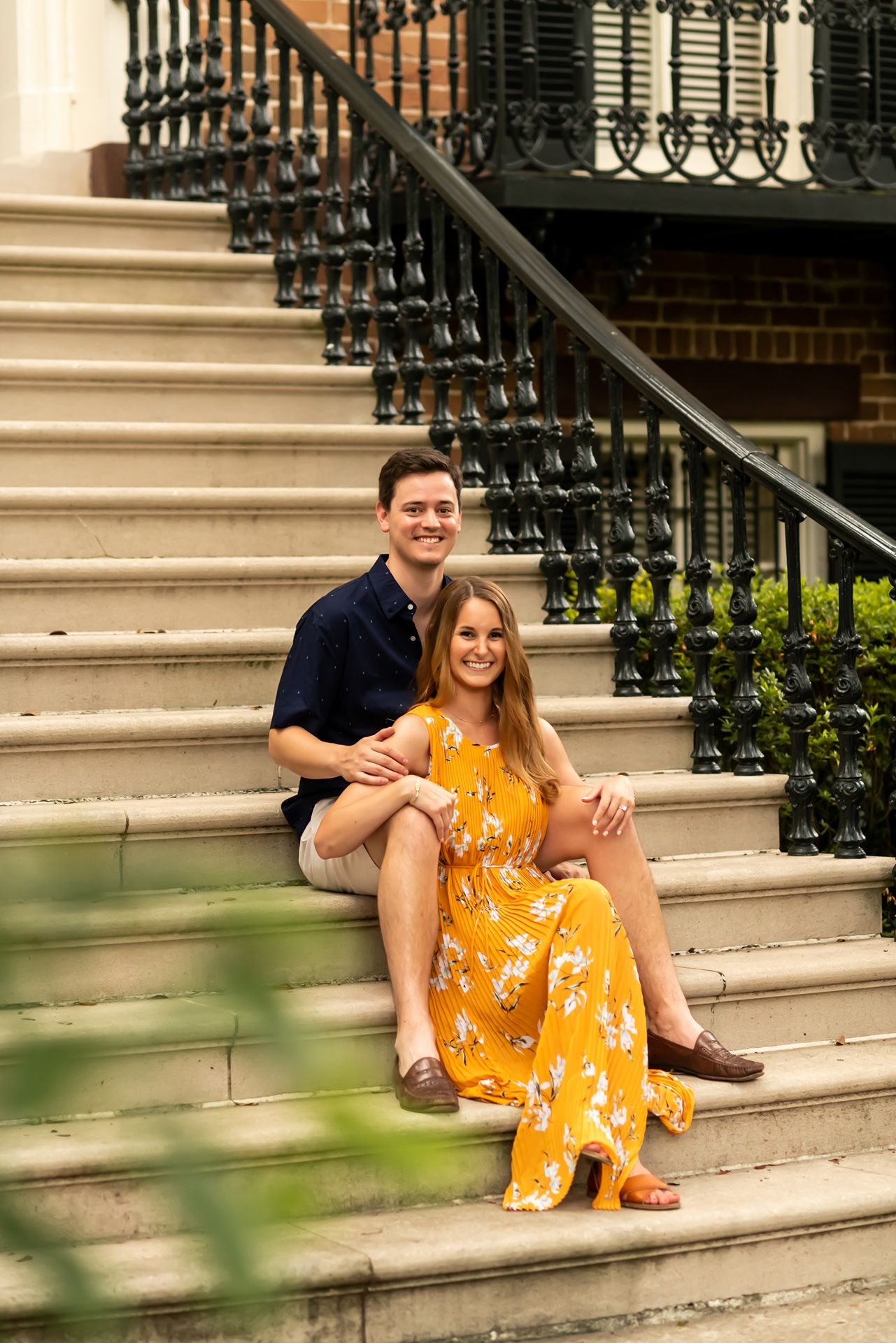 A couple sits together on outdoor stone steps with decorative black iron railings on a sunny day.