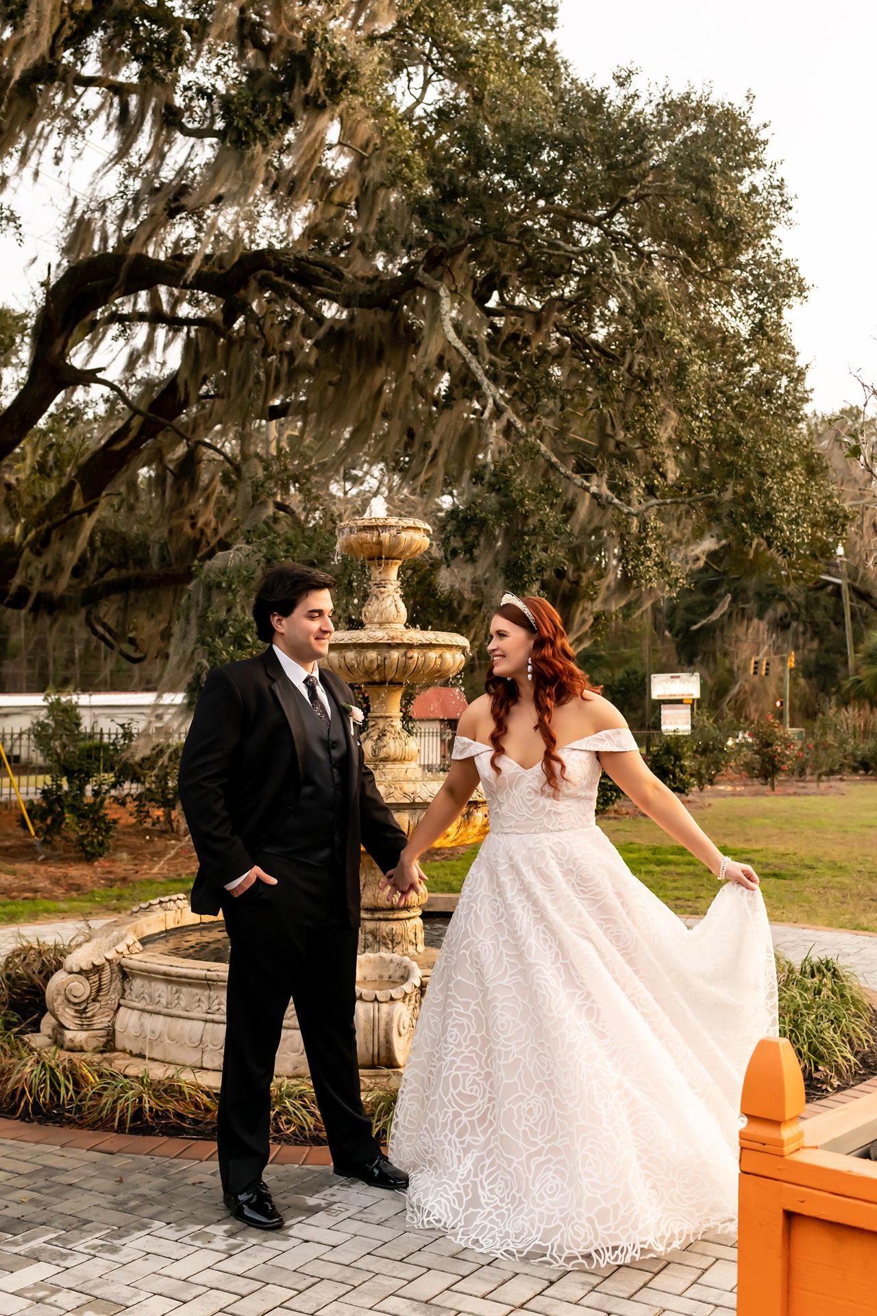 A couple poses near an ornate stone fountain in a garden atmosphere with Spanish moss draped oak trees.