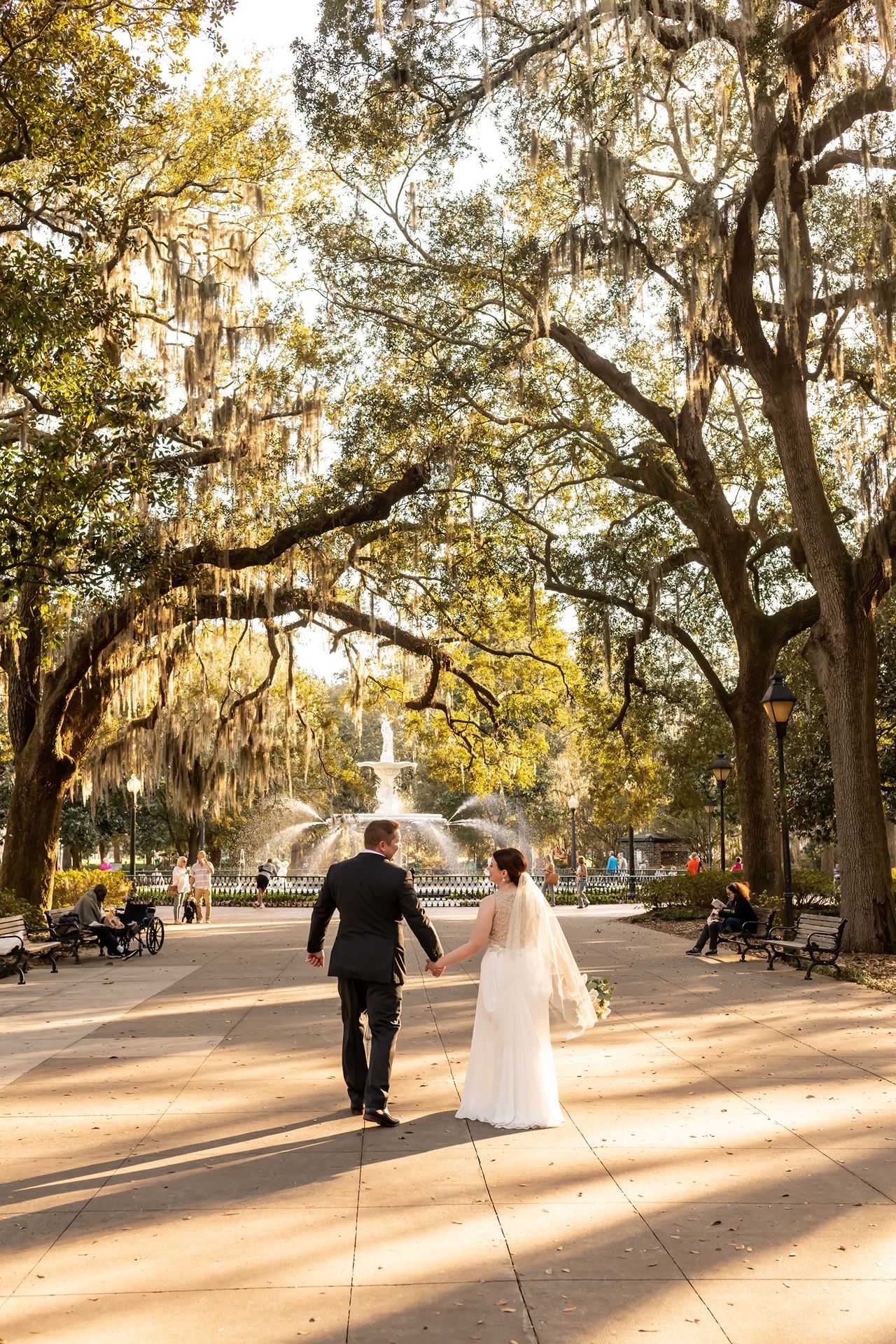A couple walking down a tree-lined path towards a fountain in a romantic Southern park with Spanish moss.