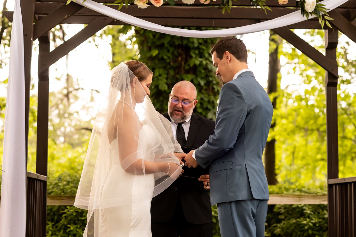A wedding ceremony takes place under a wooden pergola decorated with white fabric during a sunlit outdoor service.