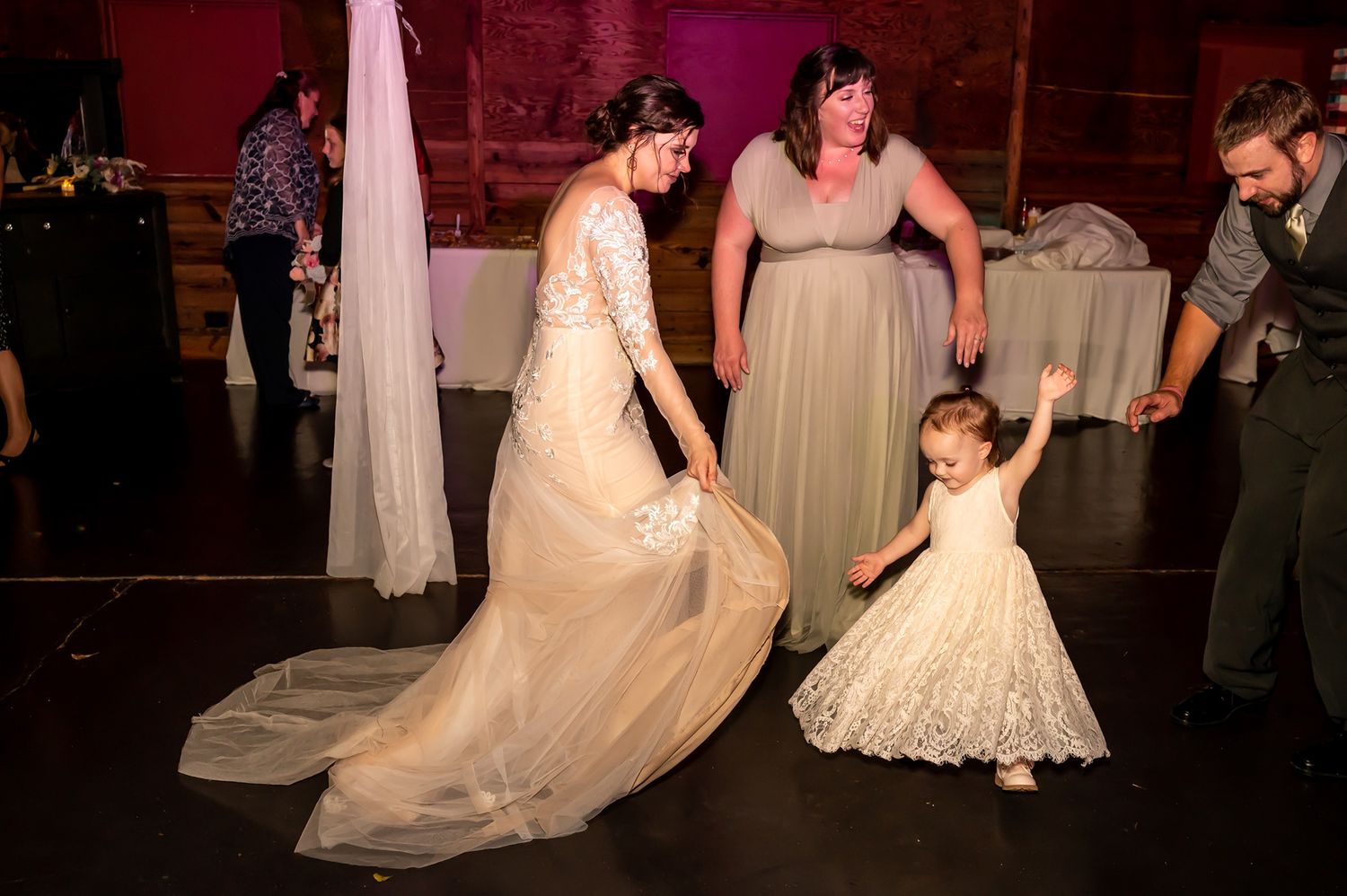 Joyful moment at a wedding reception as people dance together on a dark wooden dance floor under soft lighting.