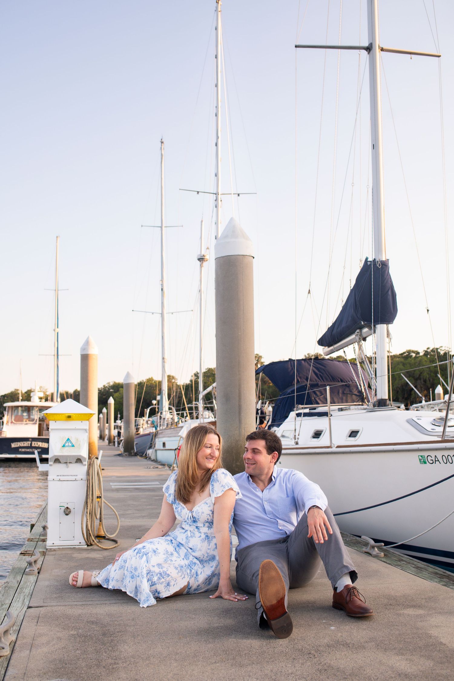 Two people share an intimate moment sitting on a marina dock surrounded by sailboats on a sunny afternoon.