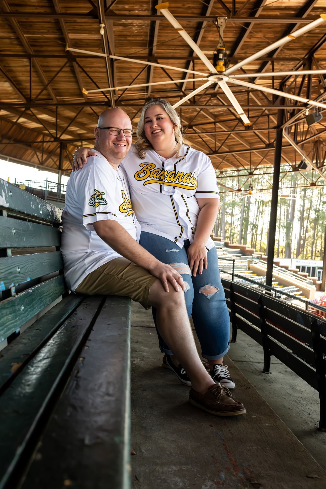 Couple wearing matching baseball jerseys sits together on green wooden bleachers under a wooden pavilion roof.