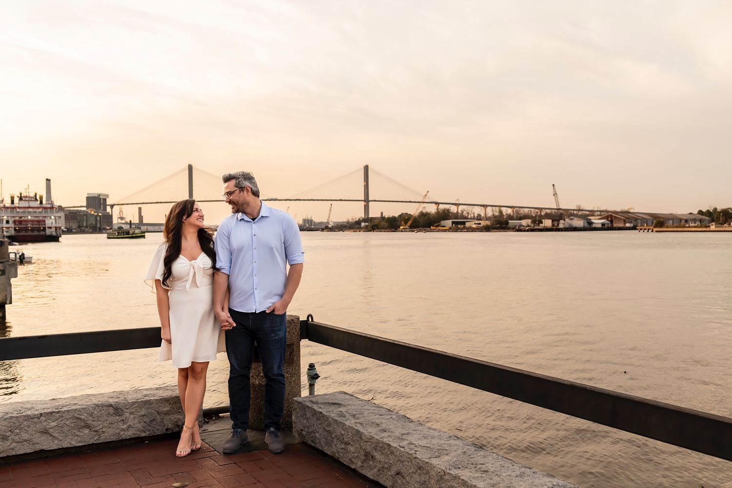 A couple stands together on a waterfront promenade with a large bridge visible across the river at sunset.