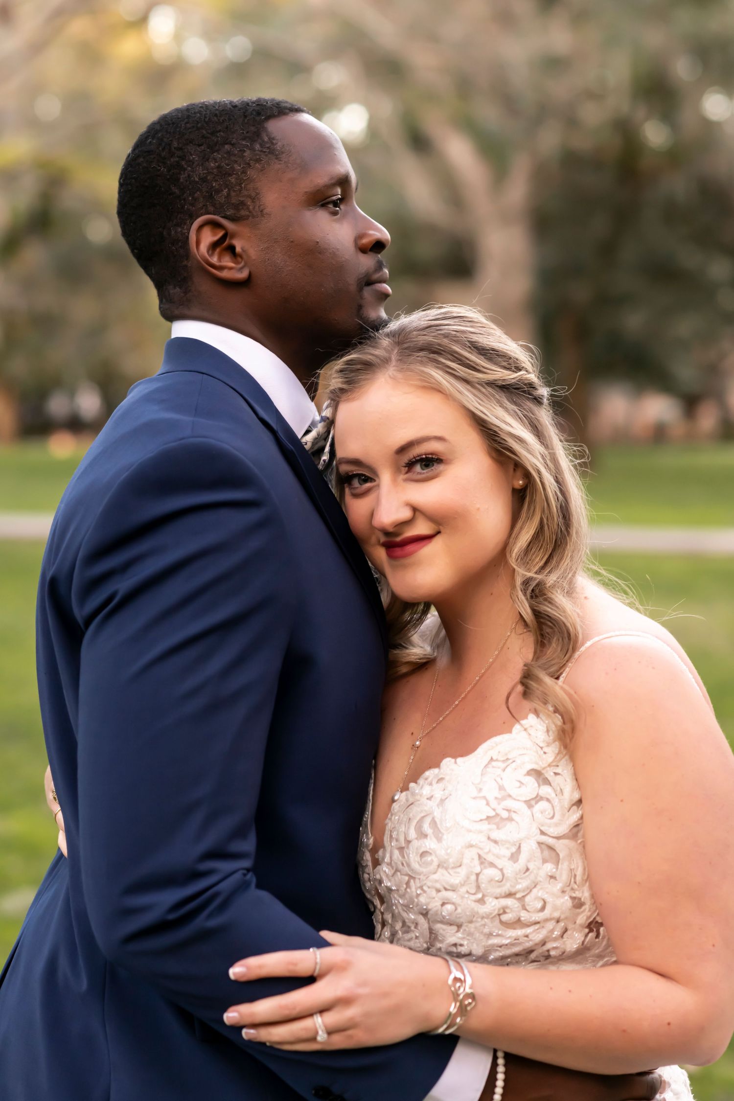 A happy bride in a white lace dress embraces her groom wearing a navy suit in a sunny outdoor setting.