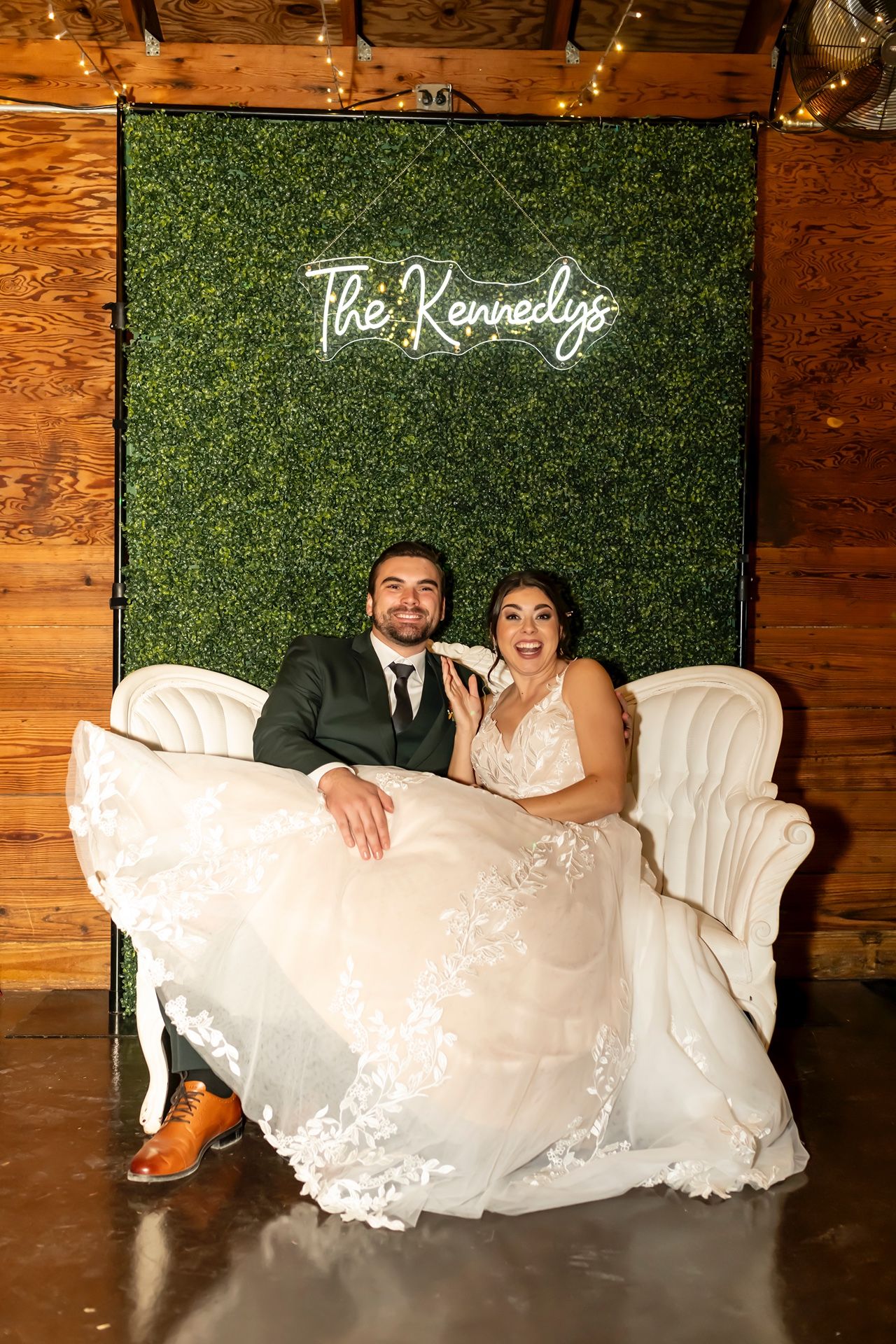 A couple lounges together on an elegant white couch against a greenery backdrop at their wedding.