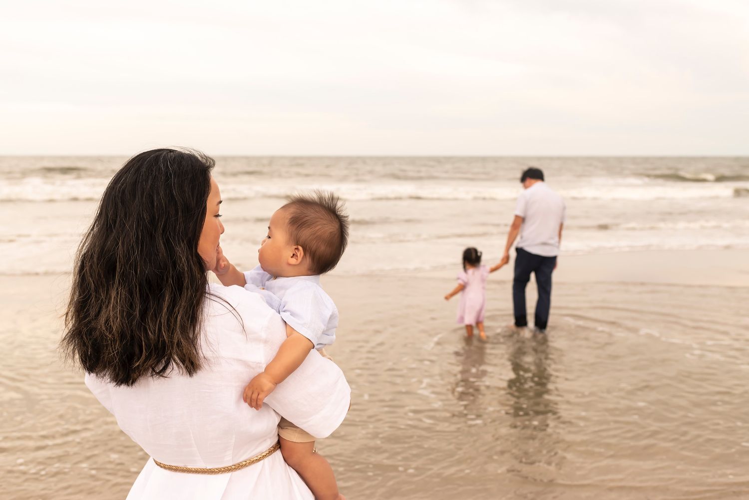 A couple and child enjoy a serene moment standing at the edge of the ocean waves on a cloudy beach day.