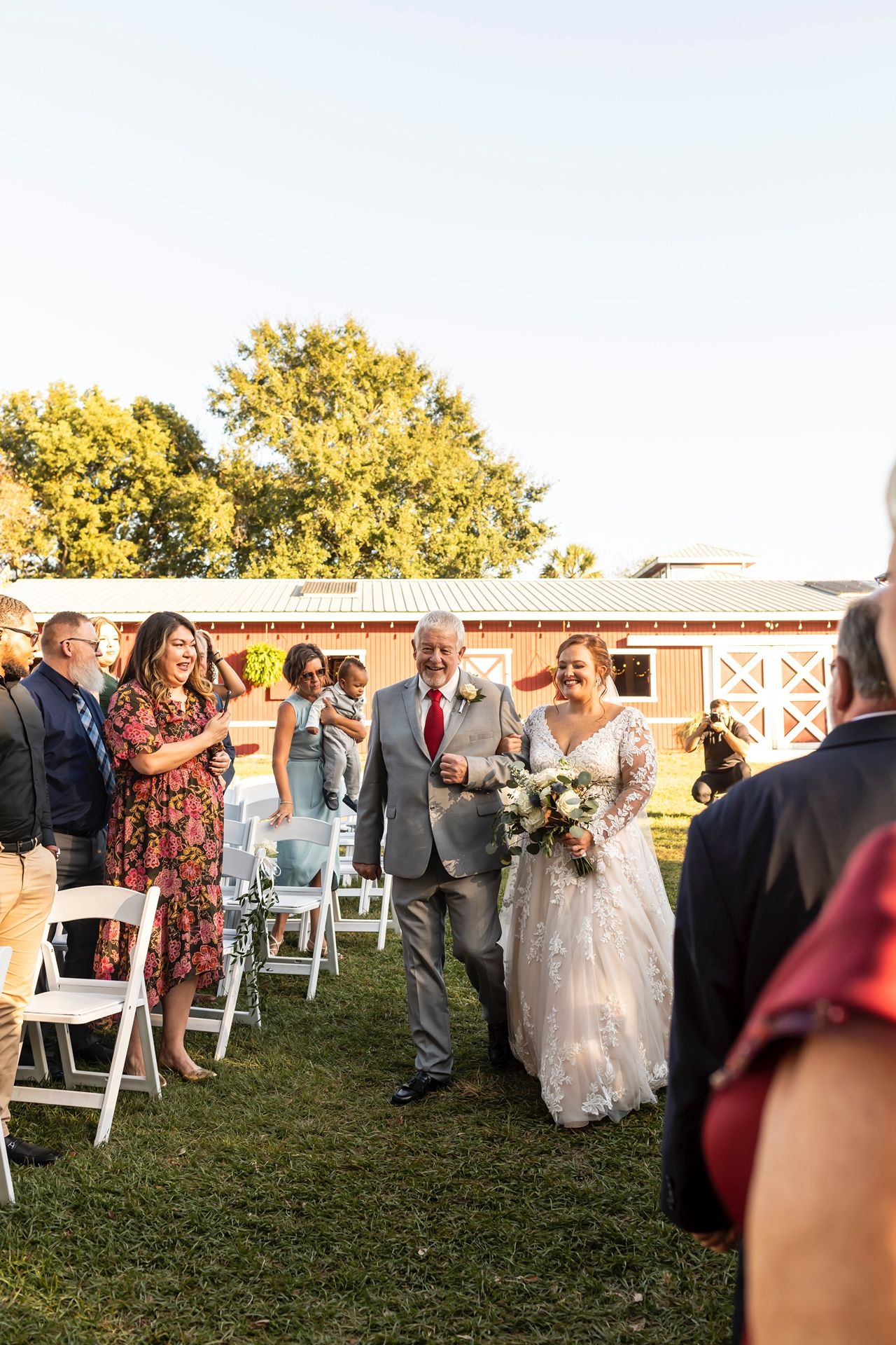 Outdoor wedding ceremony at rustic venue with guests standing on grass lawn near barn during golden hour sunset.