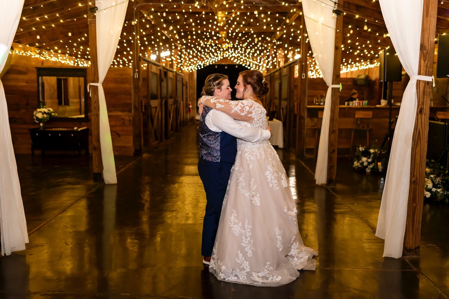 A couple embraces while dancing in a rustic venue decorated with twinkling string lights and white fabric draping.
