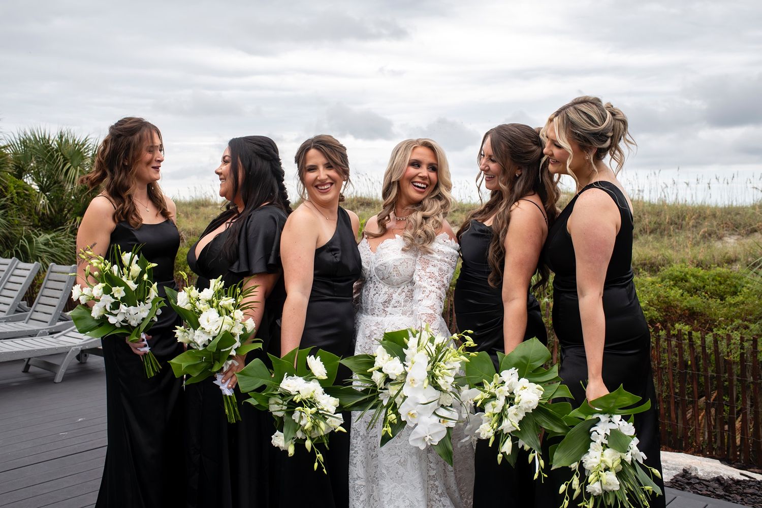 A bridal party in black dresses poses with white bouquets against a cloudy sky and natural outdoor backdrop.