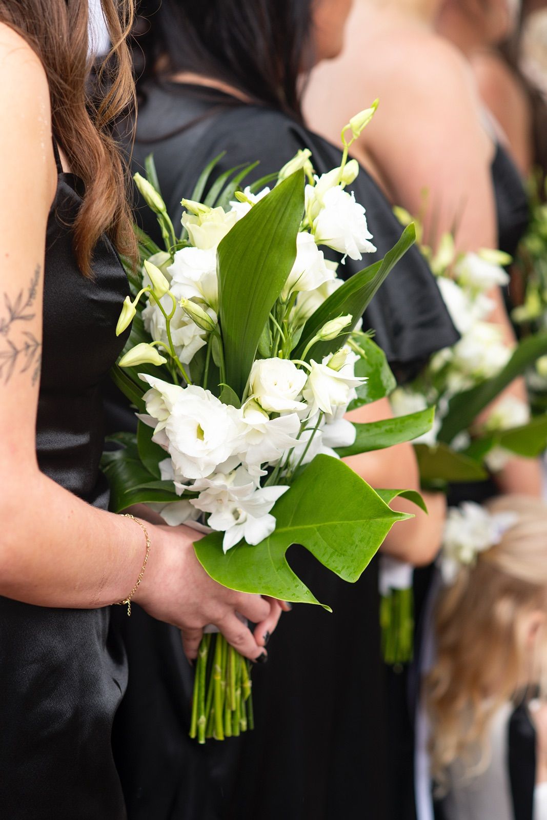 Bridesmaids in black dresses hold elegant white floral bouquets with green foliage at a formal wedding ceremony.