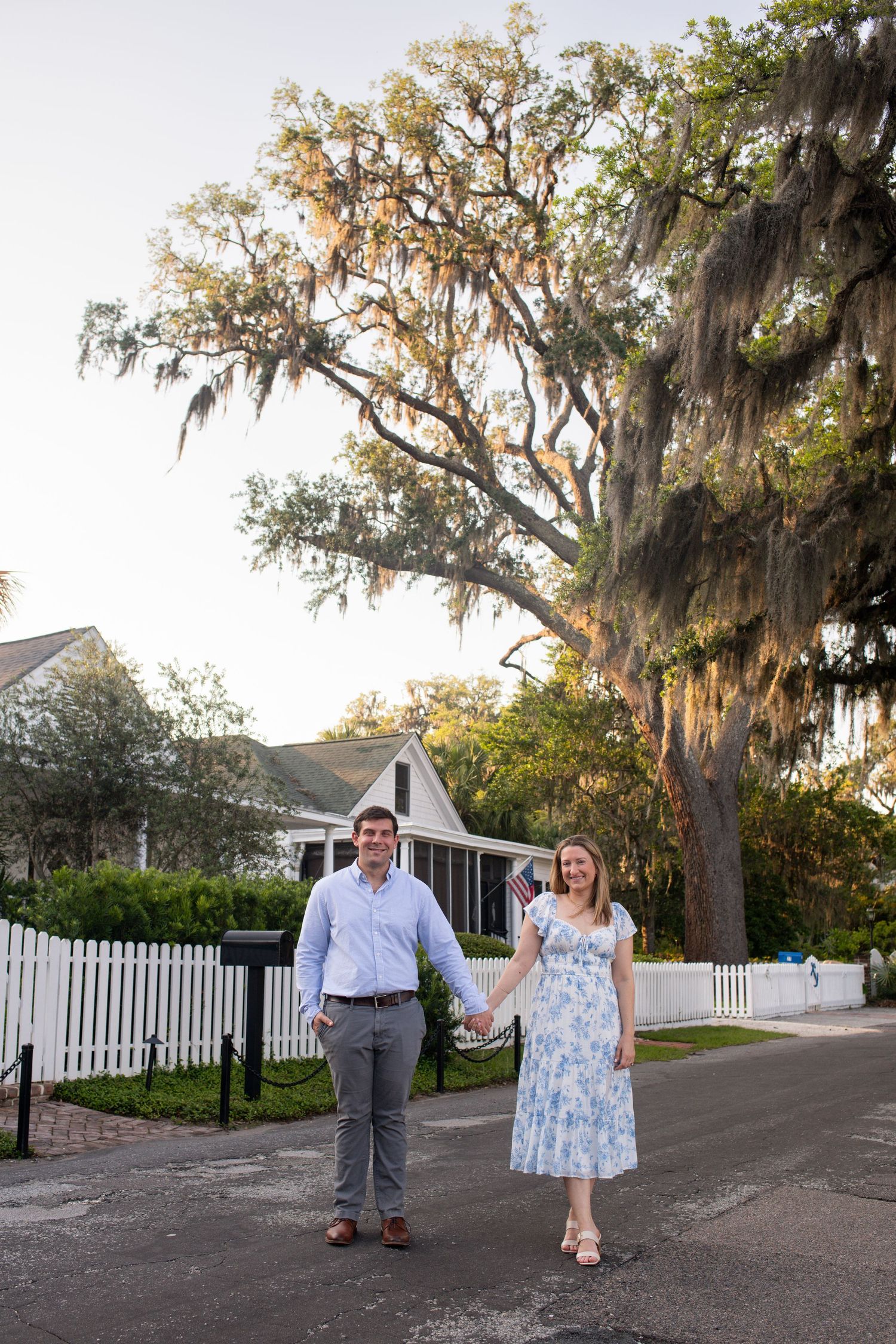 A couple walks along a tree-lined path with Spanish moss hanging from oak trees near a white picket fence.
