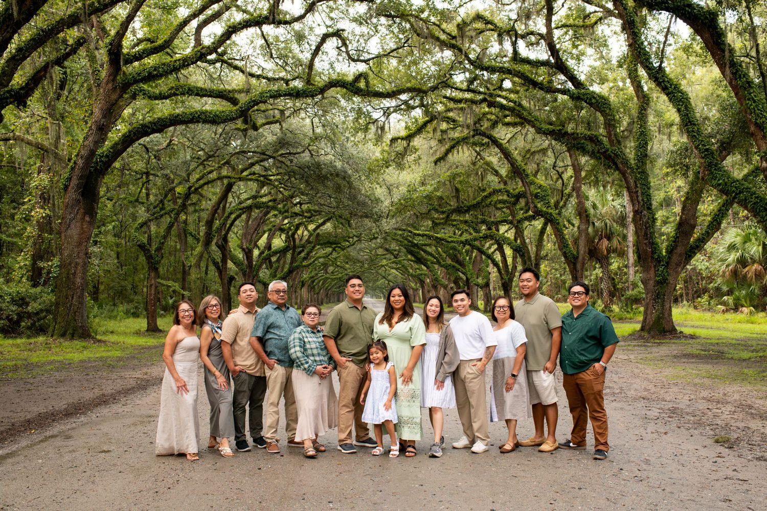 Large family group portrait under a canopy of oak trees on a scenic path in lush green surroundings.