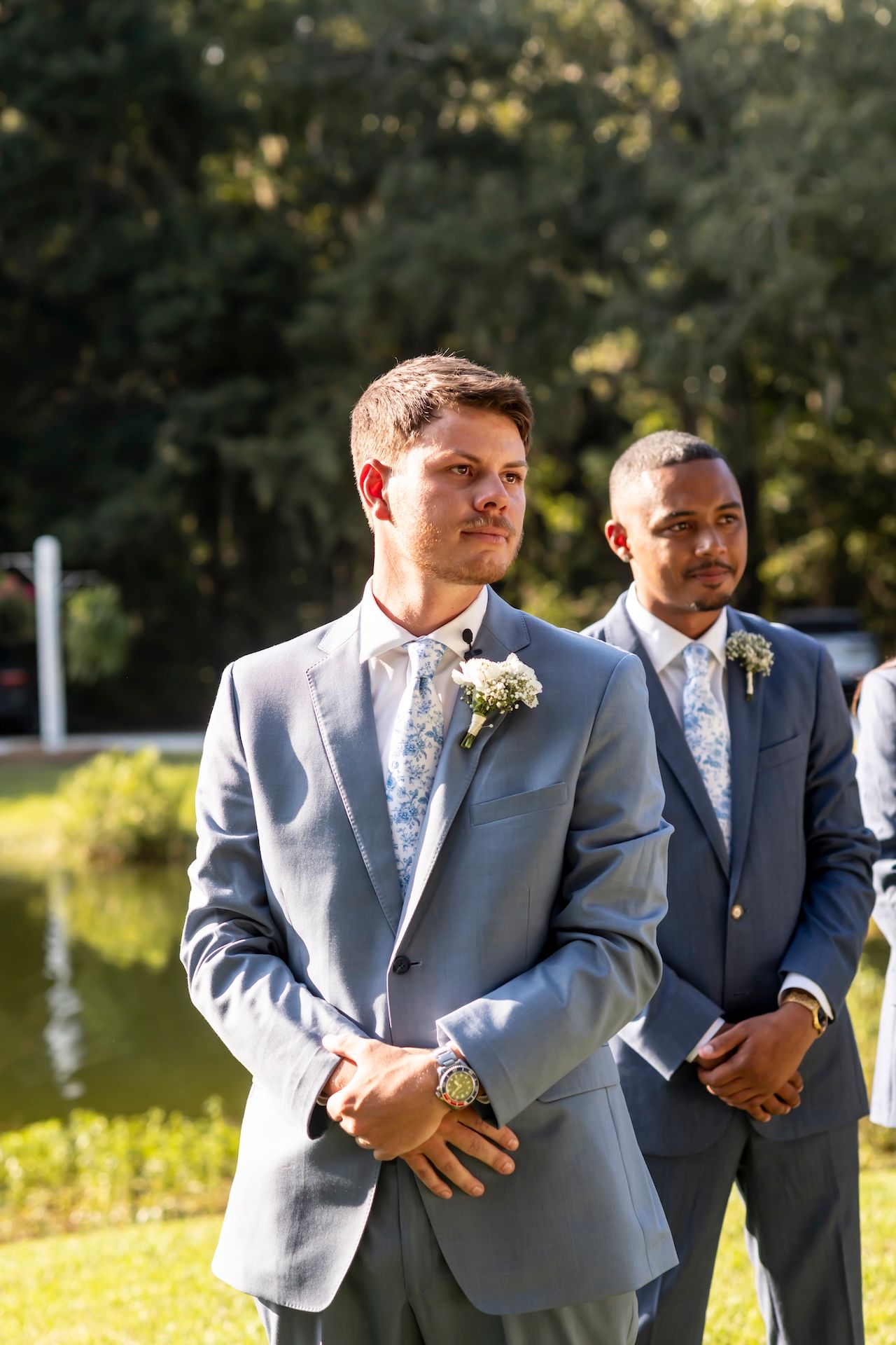 Two groomsmen in matching gray suits with floral boutonnieres stand outdoors at a wedding ceremony near a pond.