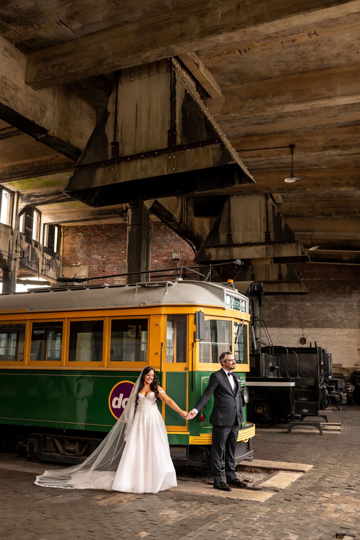 Wedding couple poses by vintage streetcar in industrial brick setting.