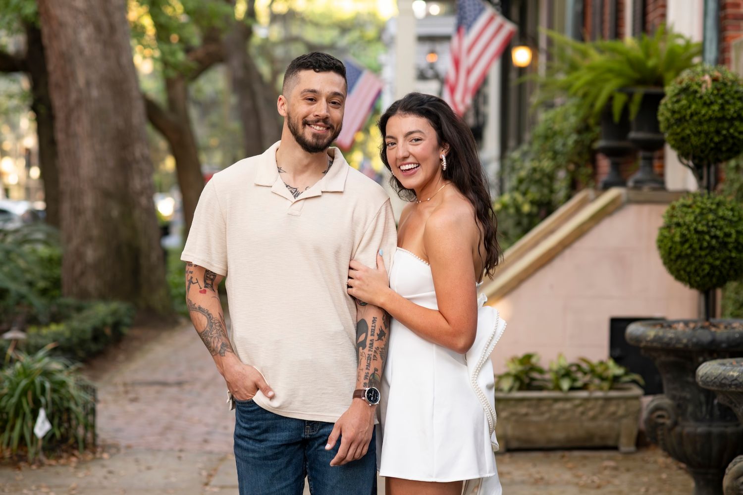 Smiling couple posing outdoors on a tree-lined sidewalk, man with tattoos in beige shirt, woman in white dress.