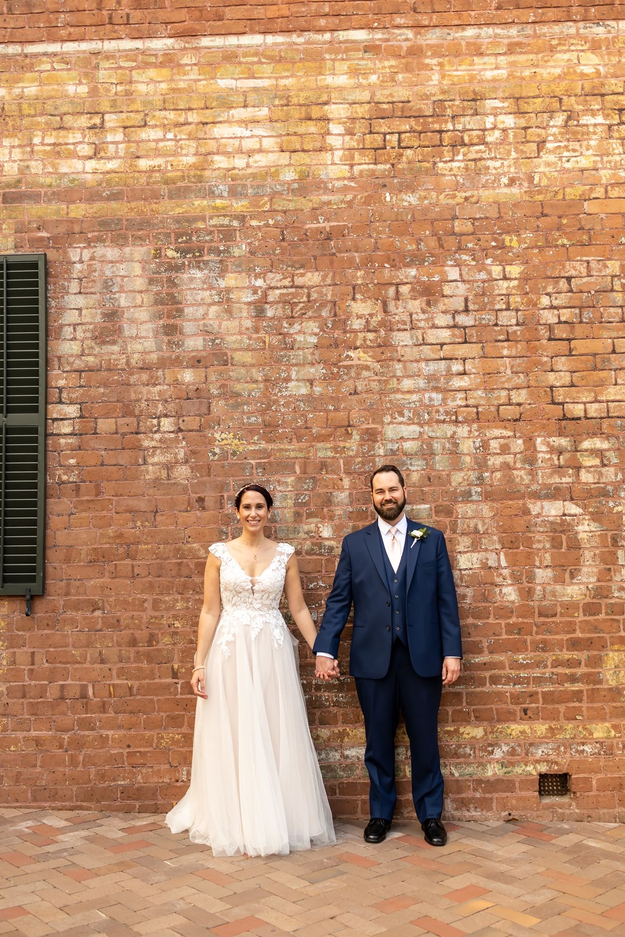 A newlywed couple stands together against a rustic brick wall at their wedding venue in formal attire.