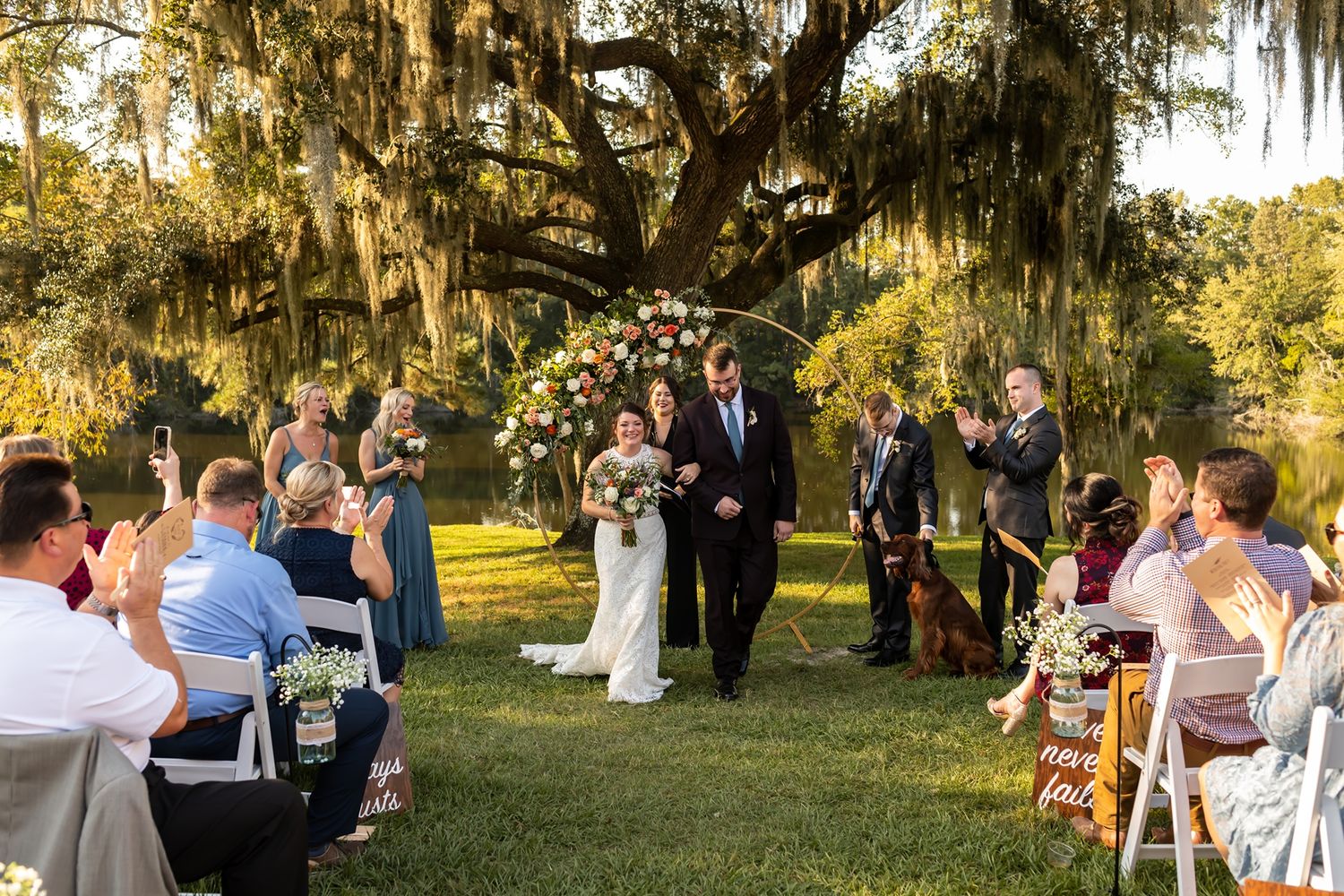 A rustic outdoor reception under live oak trees features long wooden tables and chairs set for dining at sunset.