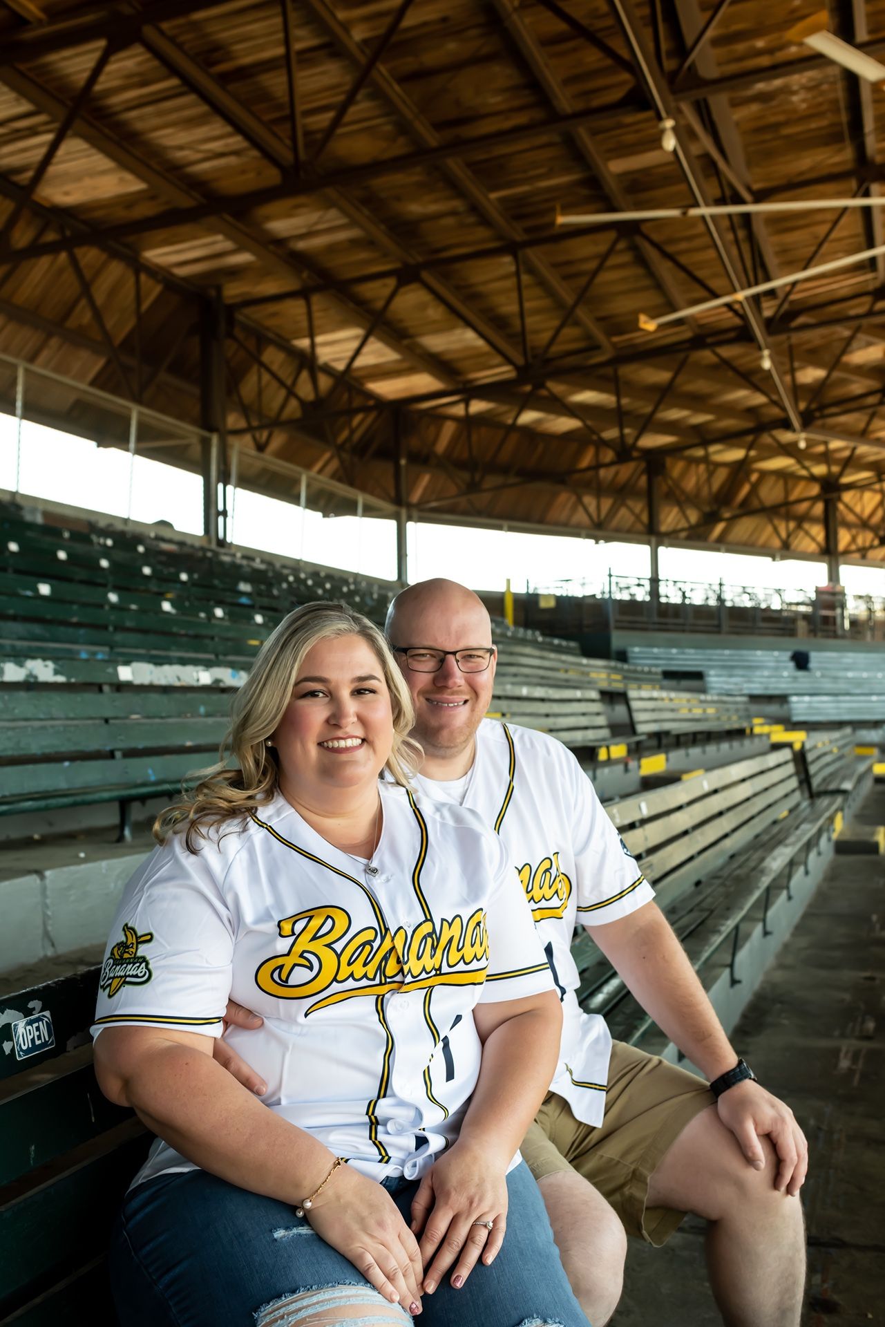 A couple wearing Savannah Bananas baseball jerseys sit together in wooden stadium bleachers under a covered roof.